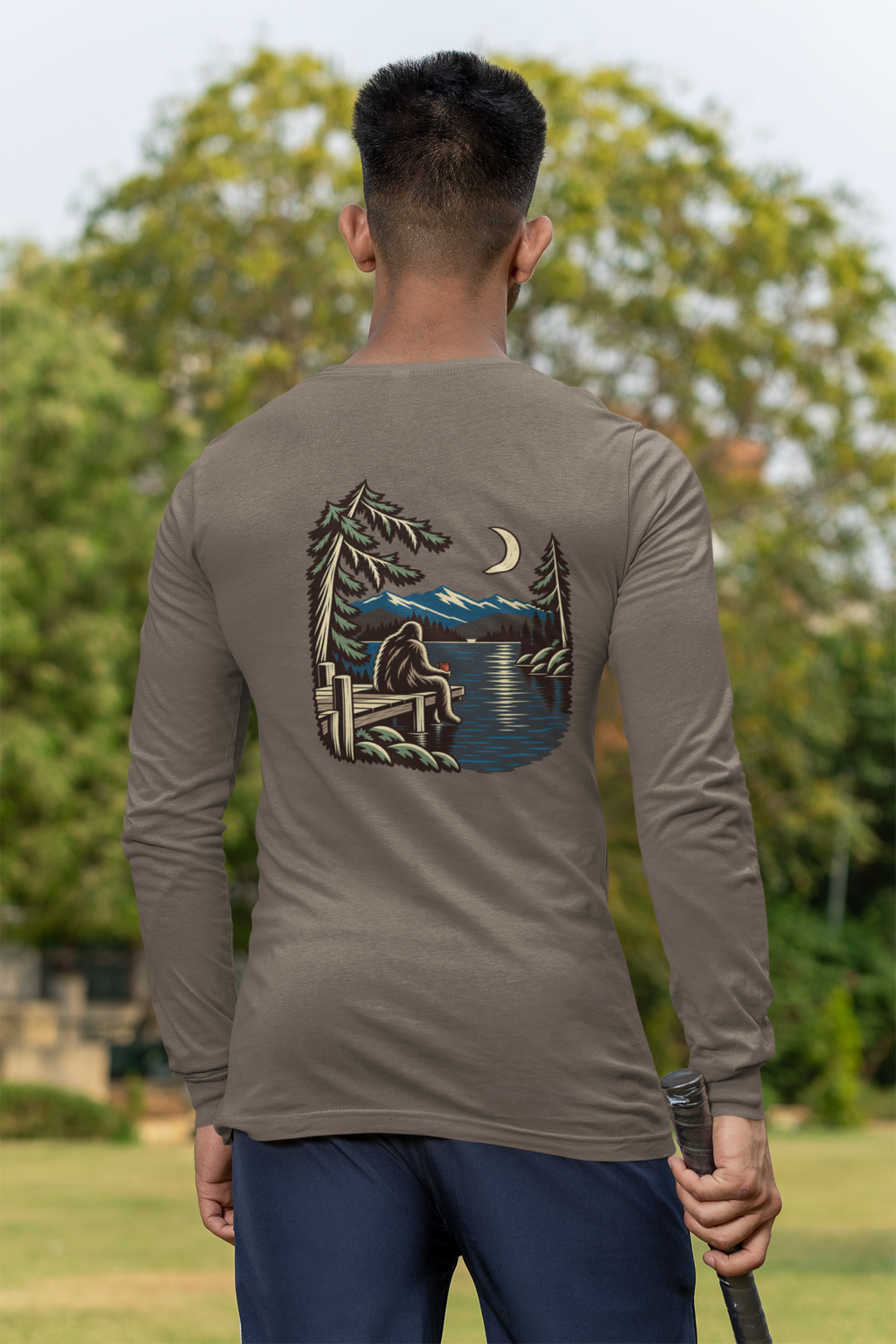 Quiet Hours Garment-Dyed Long-Sleeve Tee in Espresso, worn by a man seen from behind at a cricket field, showing the large print on the back. Digital product mockup.