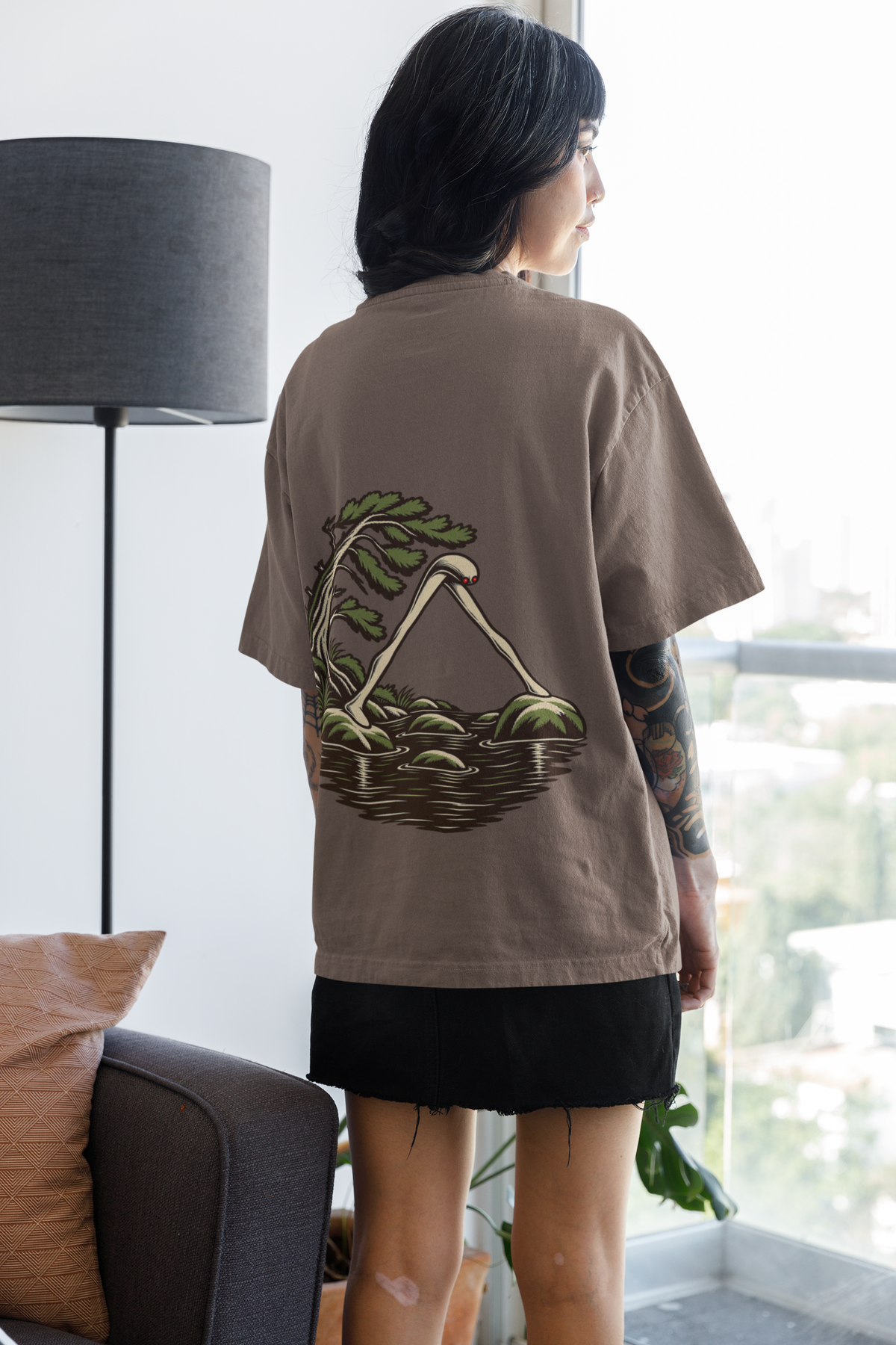 Small Hours Garment-Dyed Tee in Espresso, worn by a woman seen from behind at home by a bright window, showing the large print on the back. Digital product mockup.