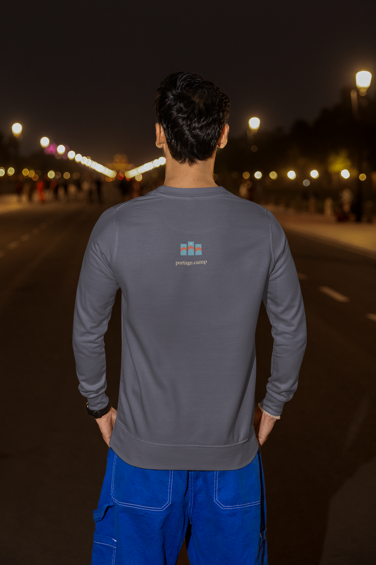 Stay for the Sky Garment-Dyed Crewneck (Front Print) in Denim, worn by a man seen from behind on a city street at night, showing the portage.camp emblem at the upper back. Digital product mockup.
