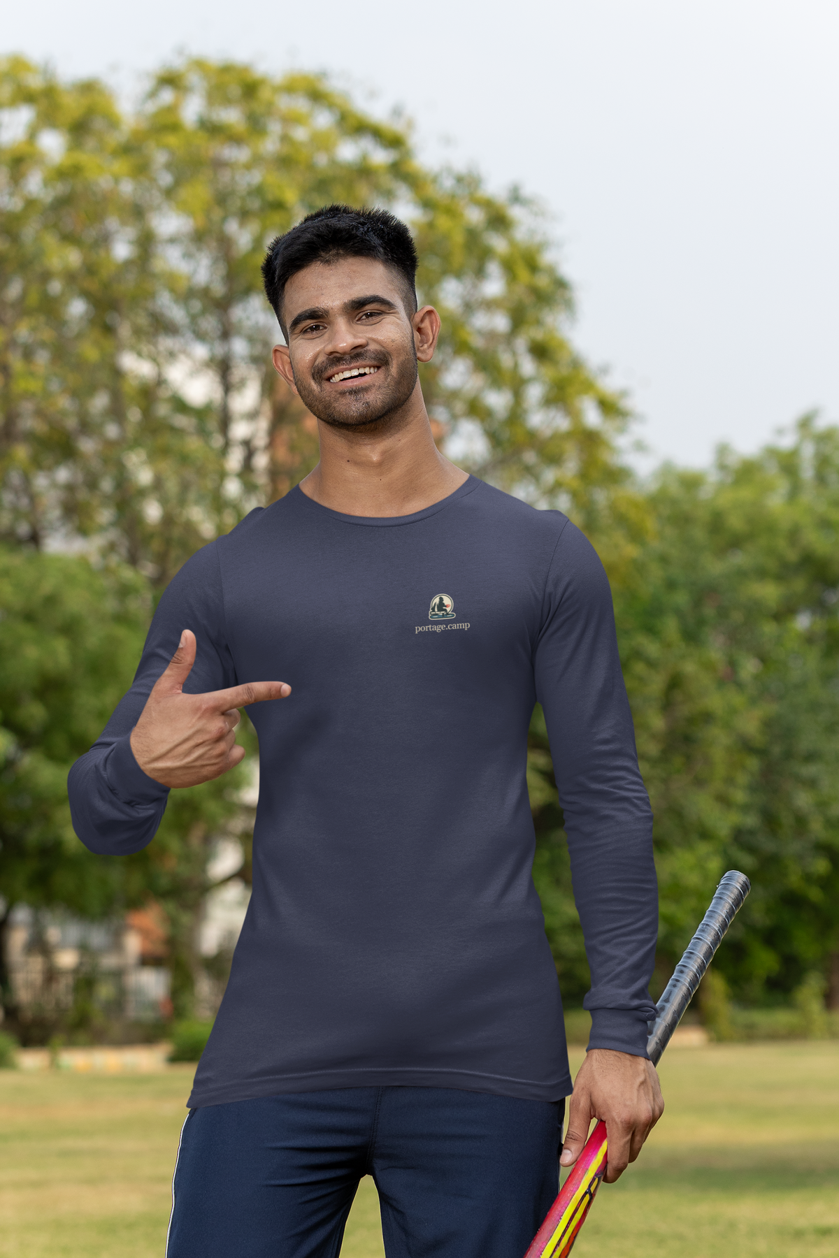 Quiet Hours Garment-Dyed Long-Sleeve Tee in True Navy, worn by a man at a cricket field, with the Quiet Hours logo and portage.camp on the chest. Digital product mockup.