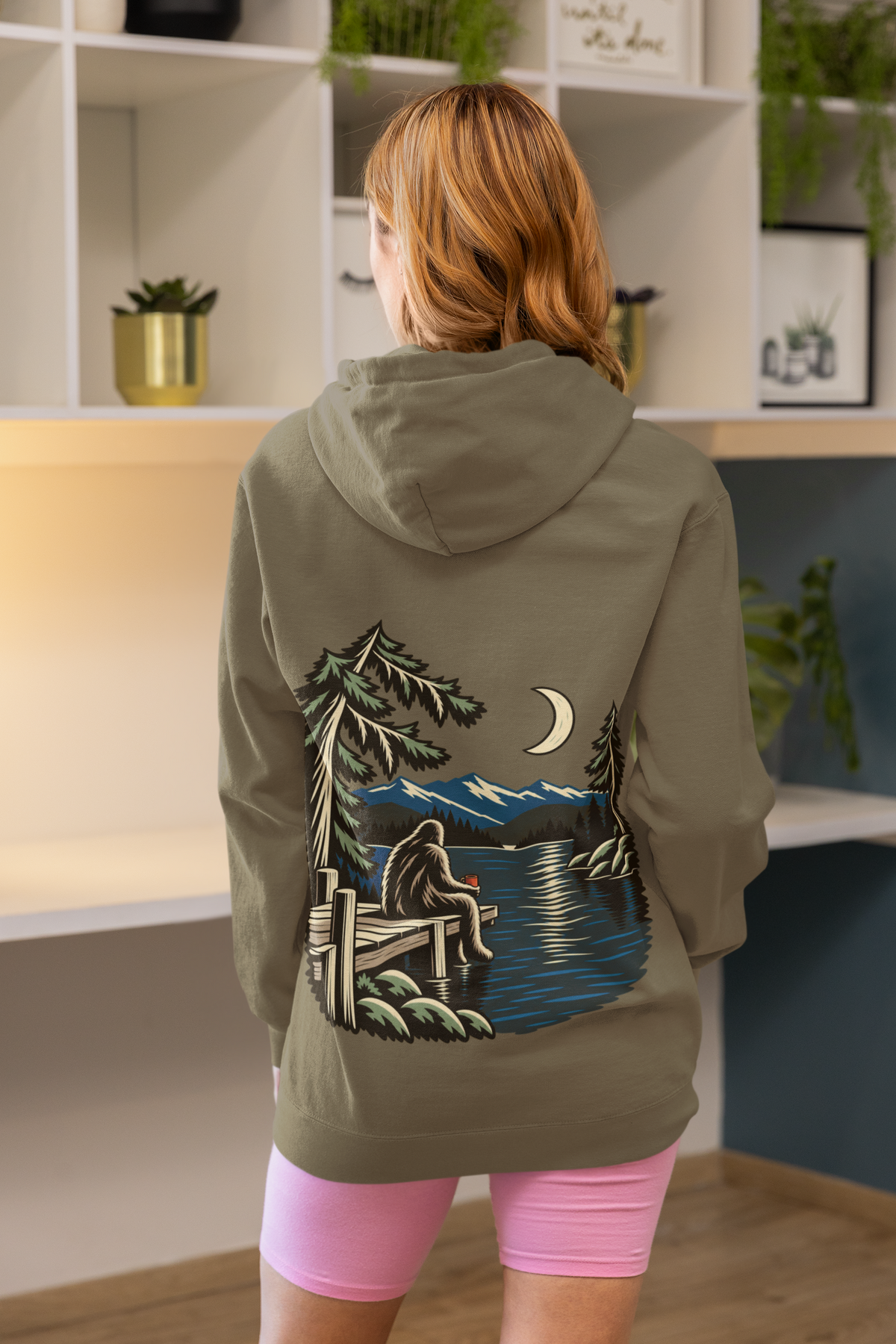 Quiet Hours Heavyweight Hoodie in Olive, worn by a woman seen from behind at home, showing the large print on the back. Digital product mockup.