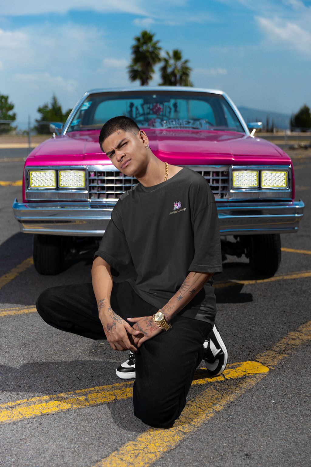 Amethyst After Hours Garment-Dyed Tee in Black, worn by a man crouched in front of a pink classic car, with the Amethyst After Hours logo and portage.camp on the chest. Digital product mockup.