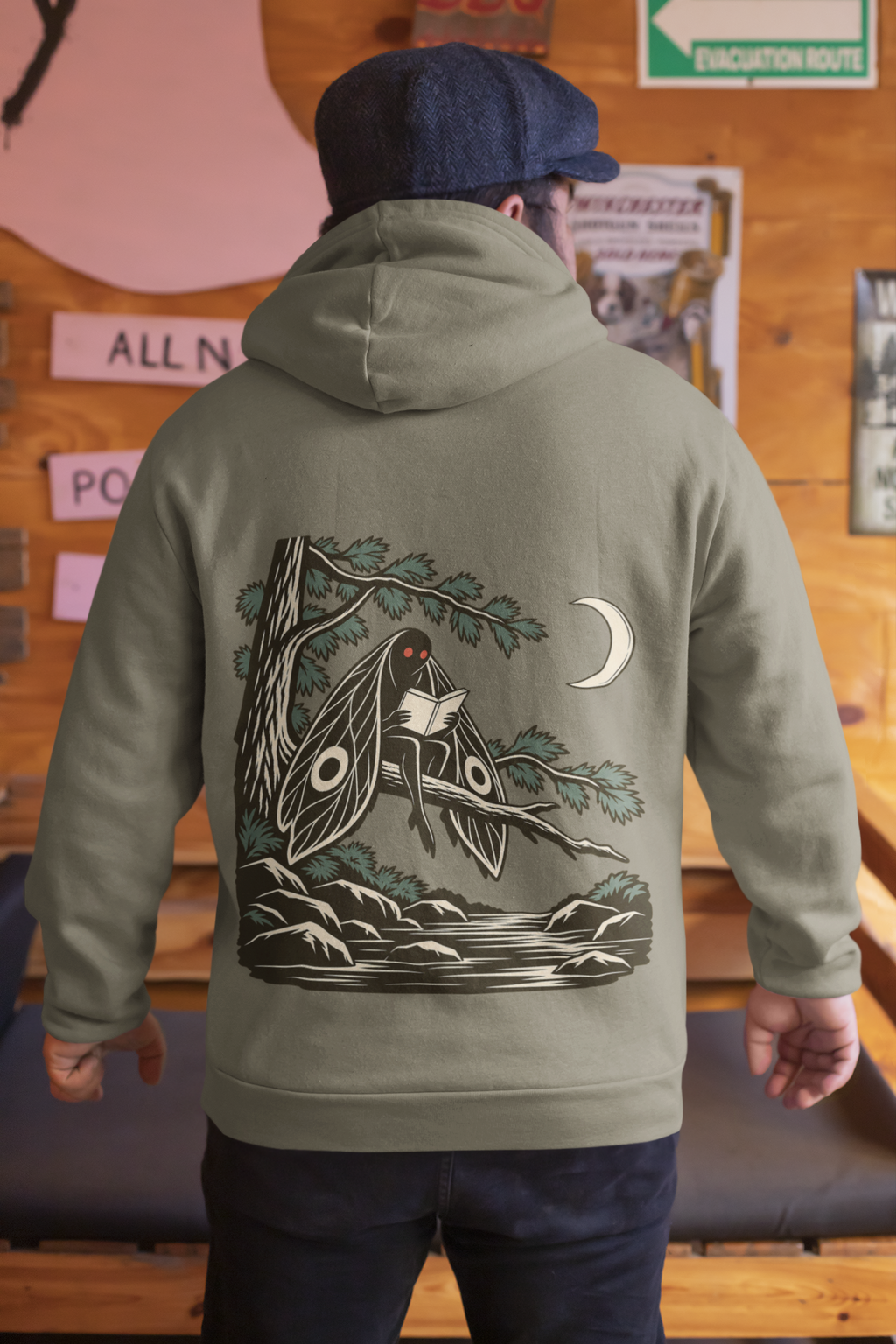 Night Library Heavyweight Hoodie in Olive, worn by a man seen from behind in a wood-panelled bar, showing the large print on the back. Digital product mockup.