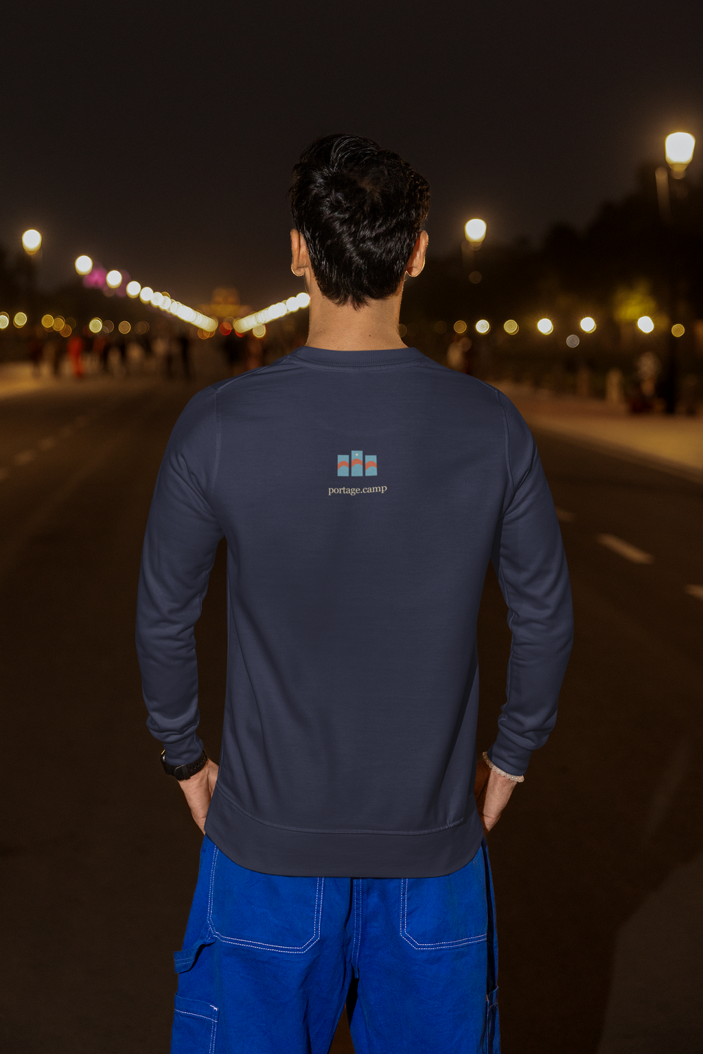 Stay for the Sky Garment-Dyed Crewneck (Front Print) in True Navy, worn by a man seen from behind on a city street at night, showing the portage.camp emblem at the upper back. Digital product mockup.