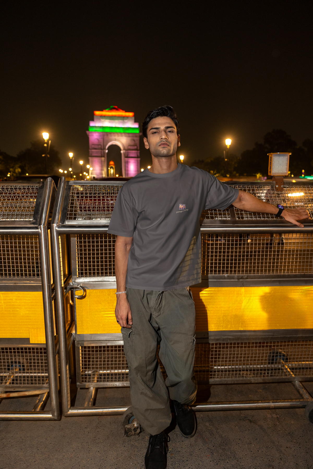 Stay for the Sky Garment-Dyed Tee in Graphite, worn by a man on a bridge over the city at night, with the Stay for the Sky logo and portage.camp on the chest. Digital product mockup.