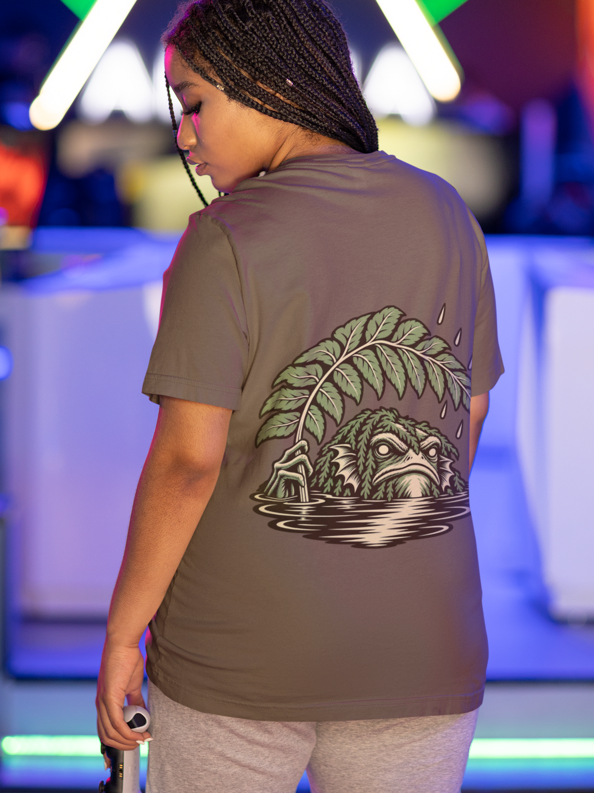 Rain Check Garment-Dyed Tee in Espresso, worn by a woman seen from behind under neon lights in a gaming arcade, showing the large print on the back. Digital product mockup.