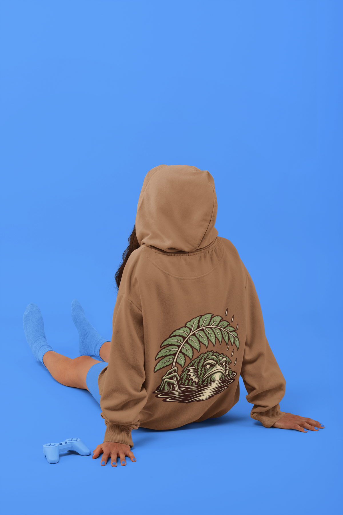 Rain Check Midweight Hoodie in Saddle, worn by a woman seen from behind seated on a studio floor, showing the large print on the back. Digital product mockup.