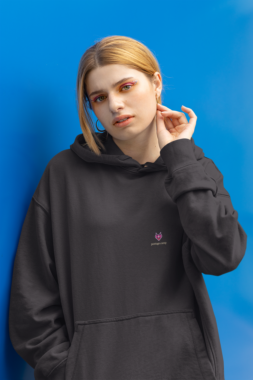 Heart Thief Midweight Hoodie in Black, worn by a woman against a bright blue wall, with the Heart Thief logo and portage.camp on the chest. Digital product mockup.