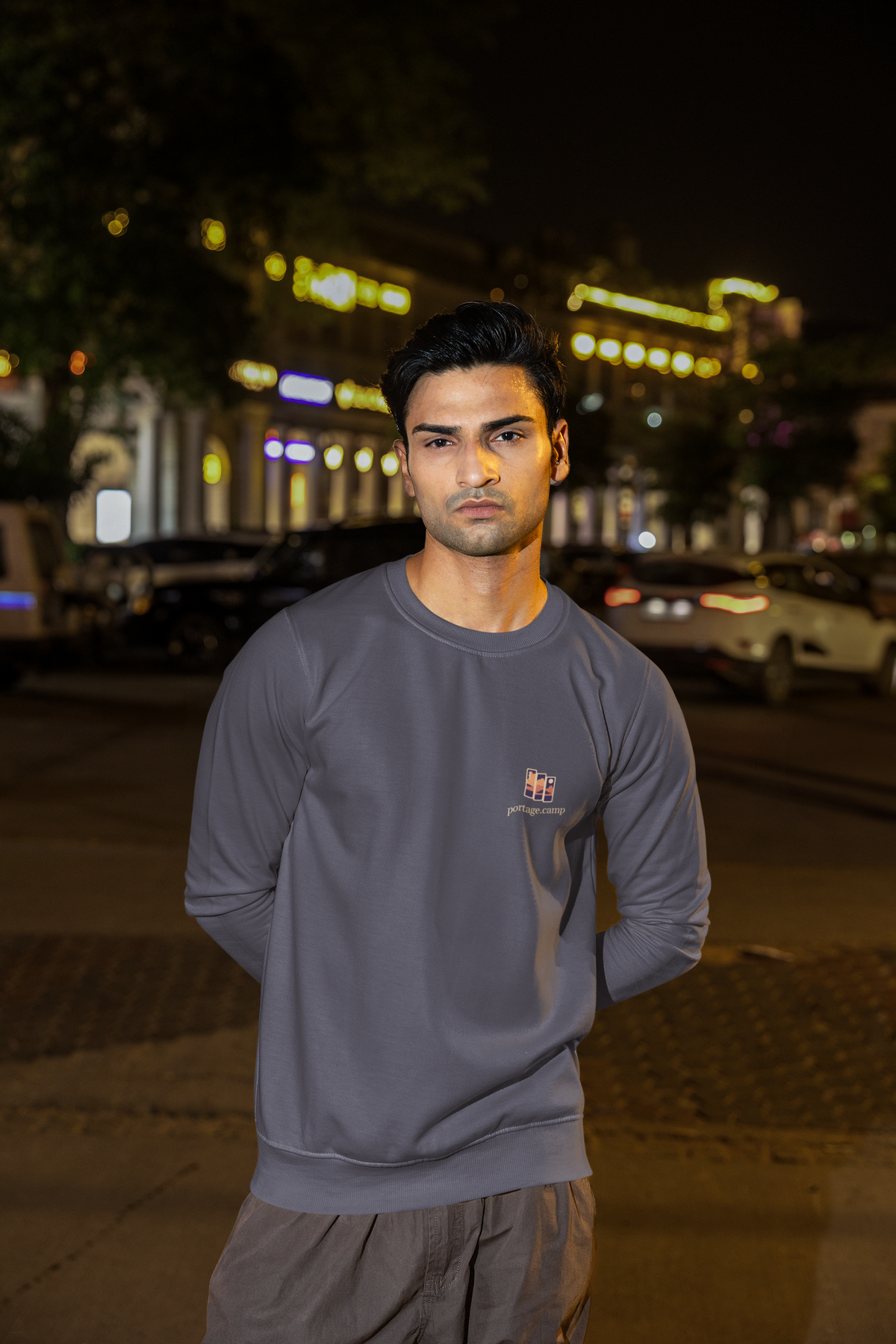 Stay for the Sky Garment-Dyed Crewneck in Denim, worn by a man on a city street at night, with the Stay for the Sky logo and portage.camp on the chest. Digital product mockup.