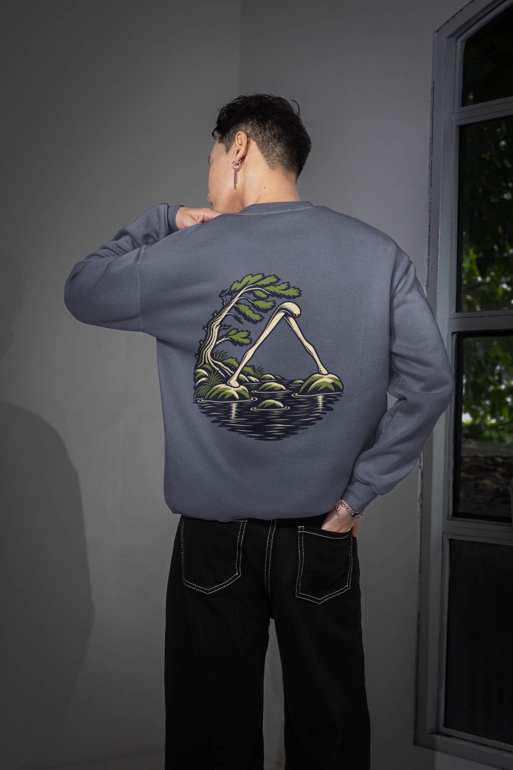 Small Hours Garment-Dyed Crewneck in Denim, worn by a man seen from behind in a room by a window, showing the large print on the back. Digital product mockup.