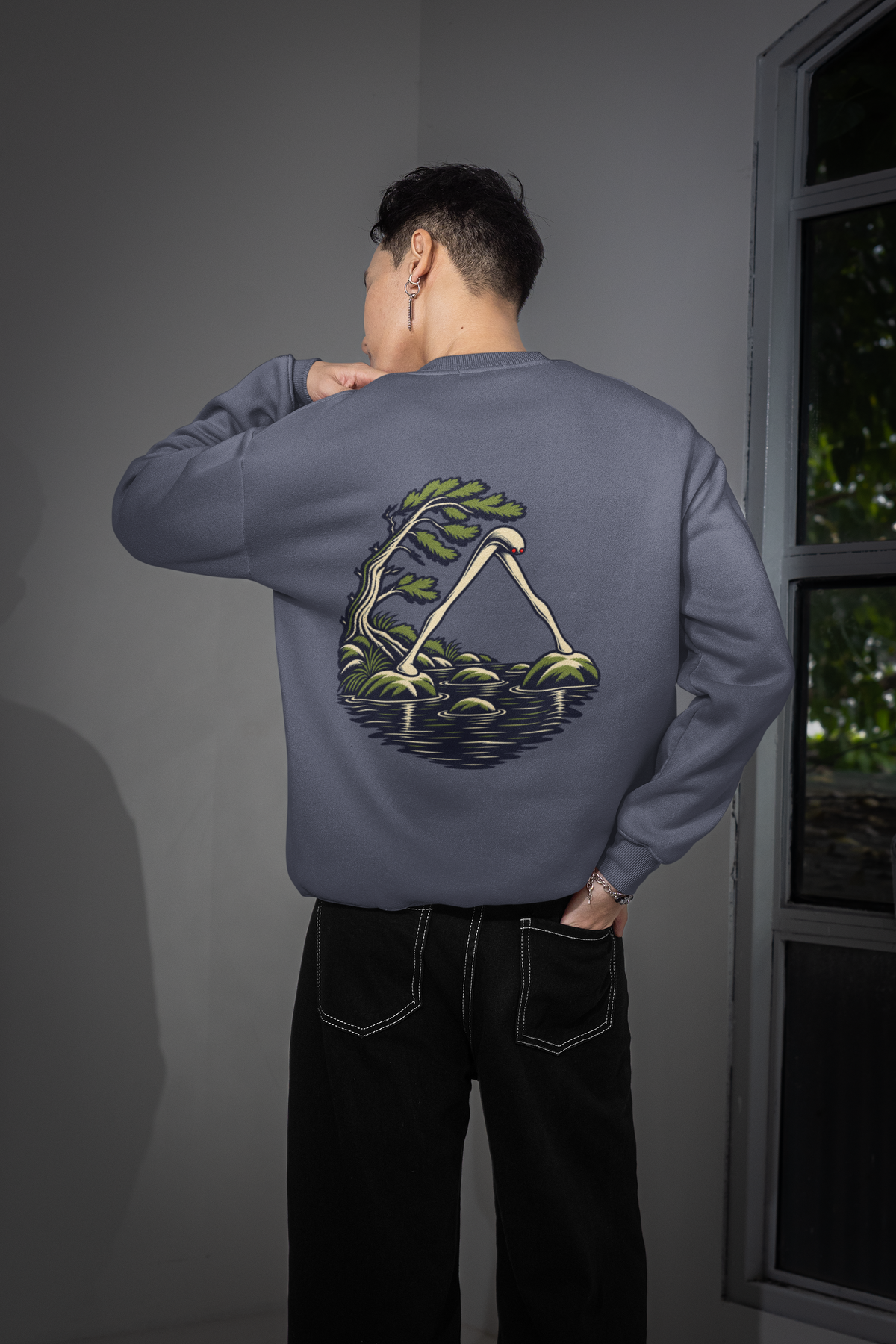 Small Hours Garment-Dyed Crewneck in Denim, worn by a man seen from behind in a room by a window, showing the large print on the back. Digital product mockup.