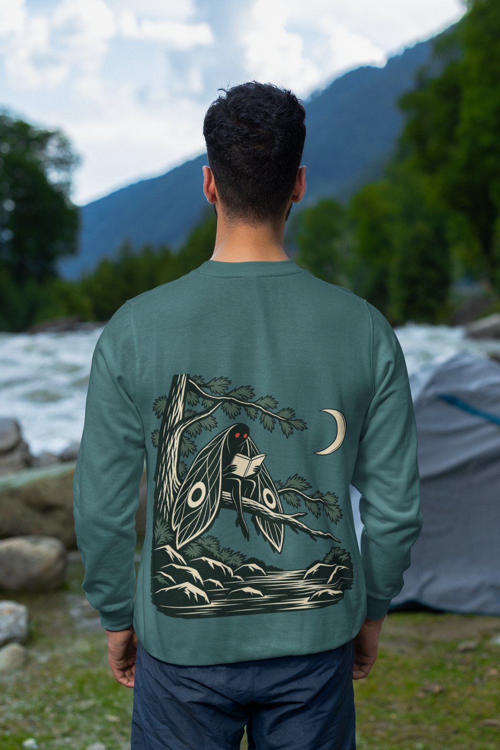 Night Library Garment-Dyed Crewneck in Blue Spruce, worn by a man seen from behind looking out over a mountain valley, showing the large print on the back. Digital product mockup.