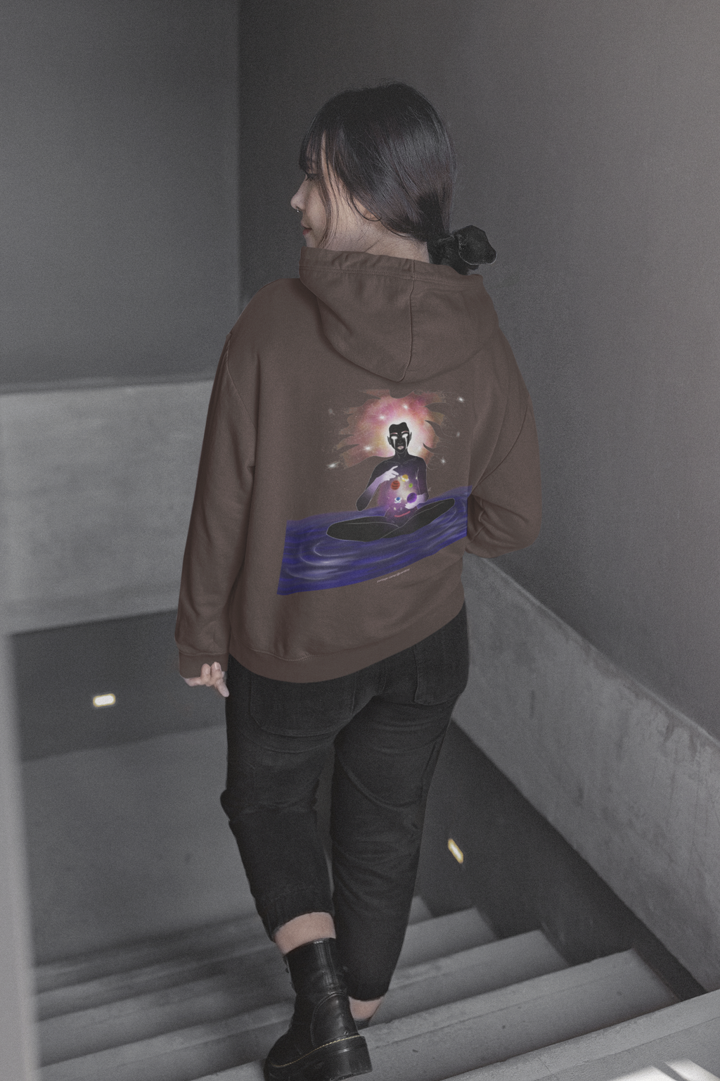 Orbit Keeper Heavyweight Hoodie in Brown, worn by a woman seen from behind walking down a staircase, showing the large print on the back. Digital product mockup.