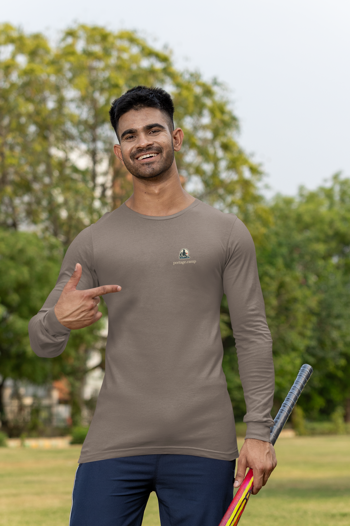 Quiet Hours Garment-Dyed Long-Sleeve Tee in Espresso, worn by a man at a cricket field, with the Quiet Hours logo and portage.camp on the chest. Digital product mockup.