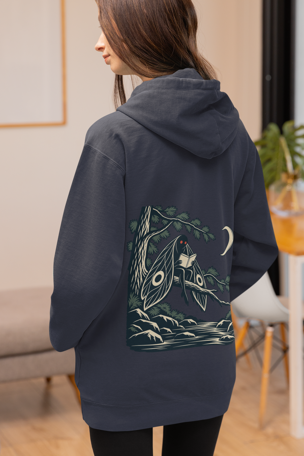 Night Library Midweight Hoodie in Classic Navy, worn by a woman seen from behind at home, showing the large print on the back. Digital product mockup.