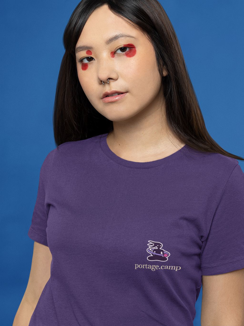 Orbit Keeper Women’s Relaxed Tee in Team Purple, worn by a woman with graphic eyeliner, close up, with the Orbit Keeper logo and portage.camp on the chest. Digital product mockup.
