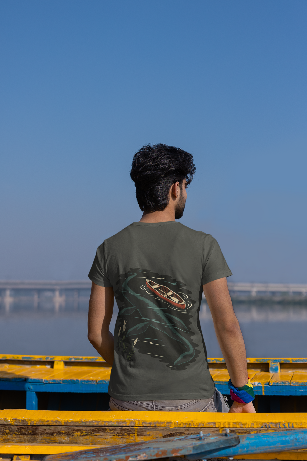 Seen Briefly Garment-Dyed Tee in Pepper, worn by a man seen from behind seated on a canoe by the water, showing the large print on the back. Digital product mockup.