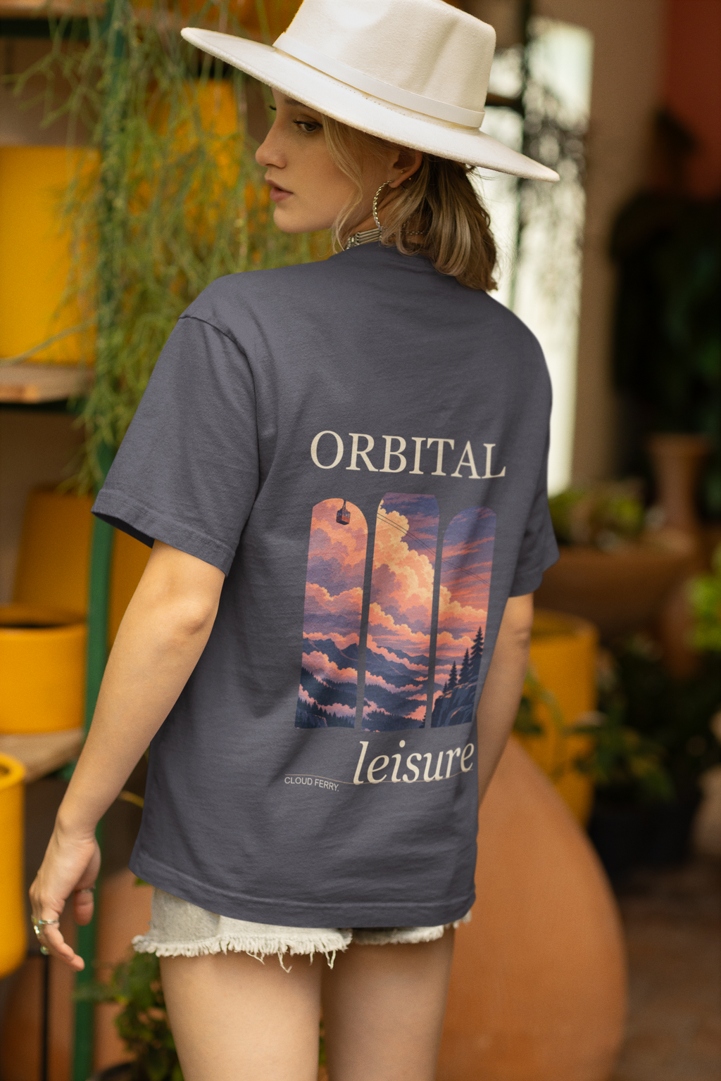 Cloud Ferry Garment-Dyed Tee in Navy, worn by a woman seen from behind in a sun hat among potted plants, showing the large print on the back. Digital product mockup.