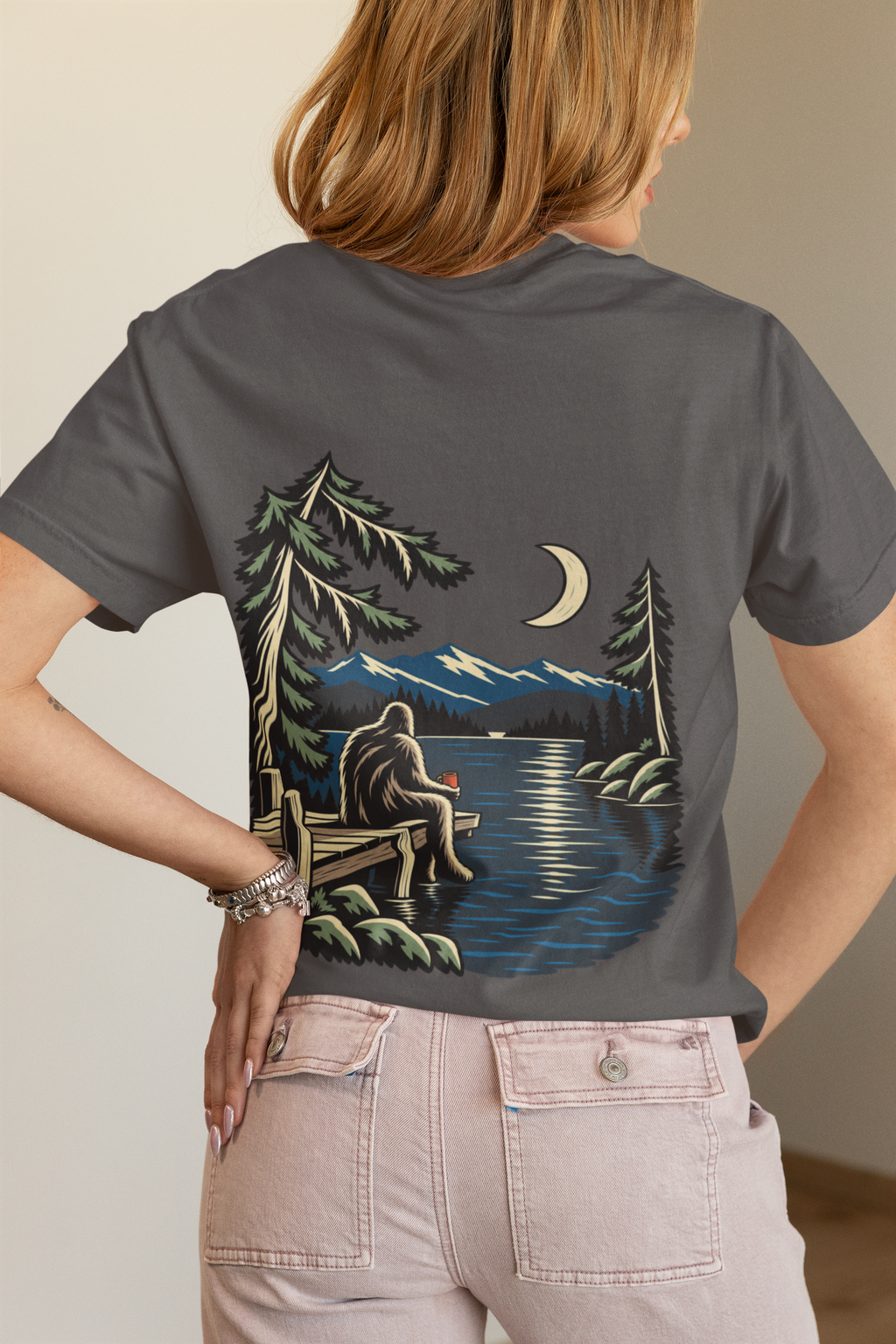 Quiet Hours Women’s Relaxed Tee in Asphalt, worn by a woman seen from behind in a bedroom at home, showing the large print on the back. Digital product mockup.