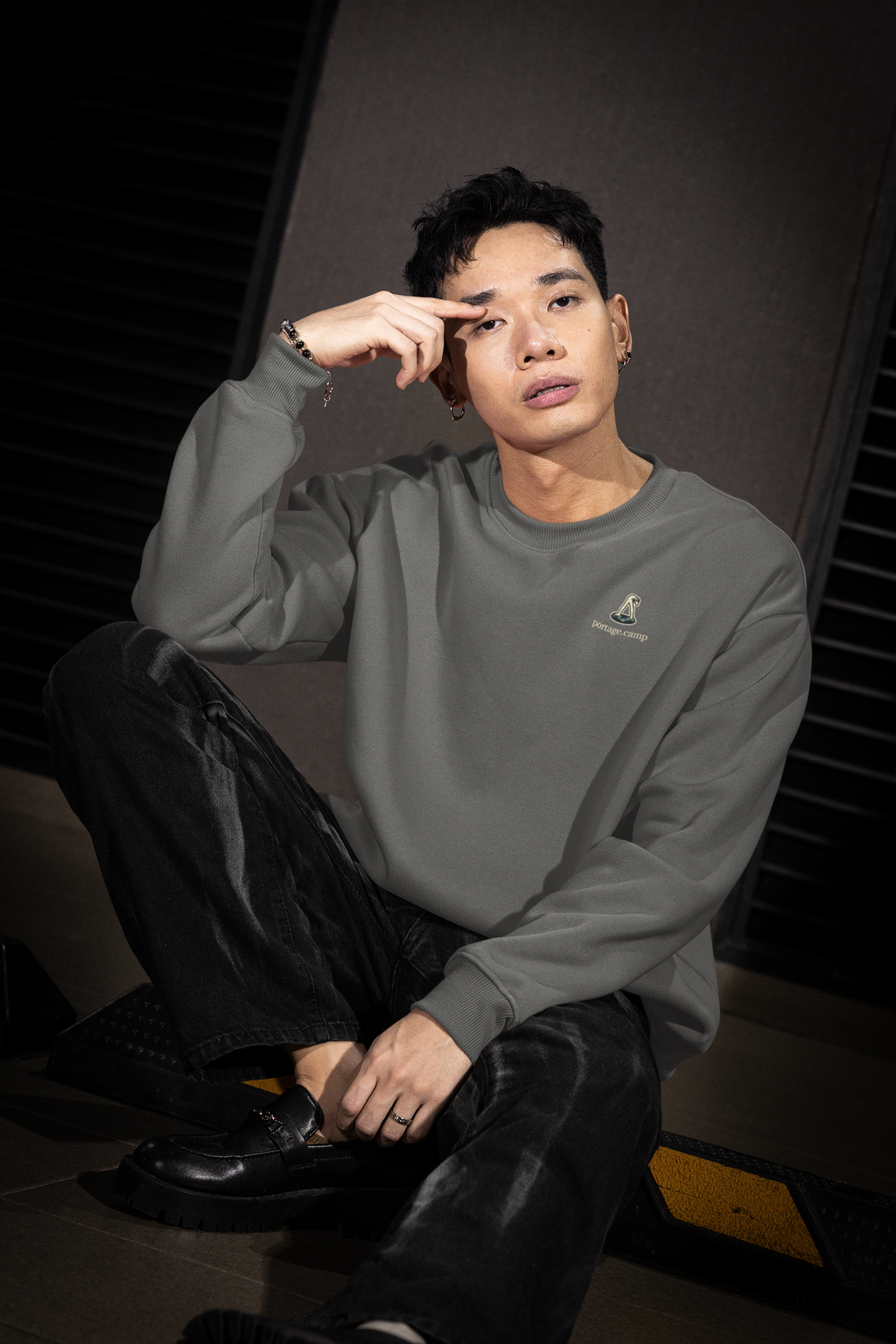Small Hours Garment-Dyed Crewneck in Pepper, worn by a man seated on a city sidewalk, with the Small Hours logo and portage.camp on the chest. Digital product mockup.