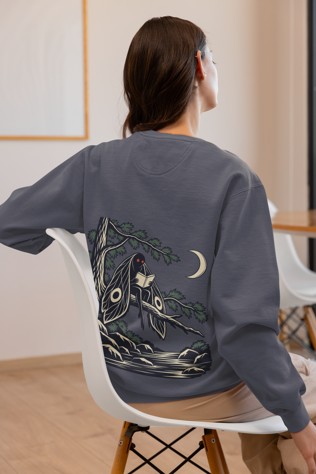 Night Library Garment-Dyed Crewneck in Denim, worn by a woman seen from behind at home, showing the large print on the back. Digital product mockup.