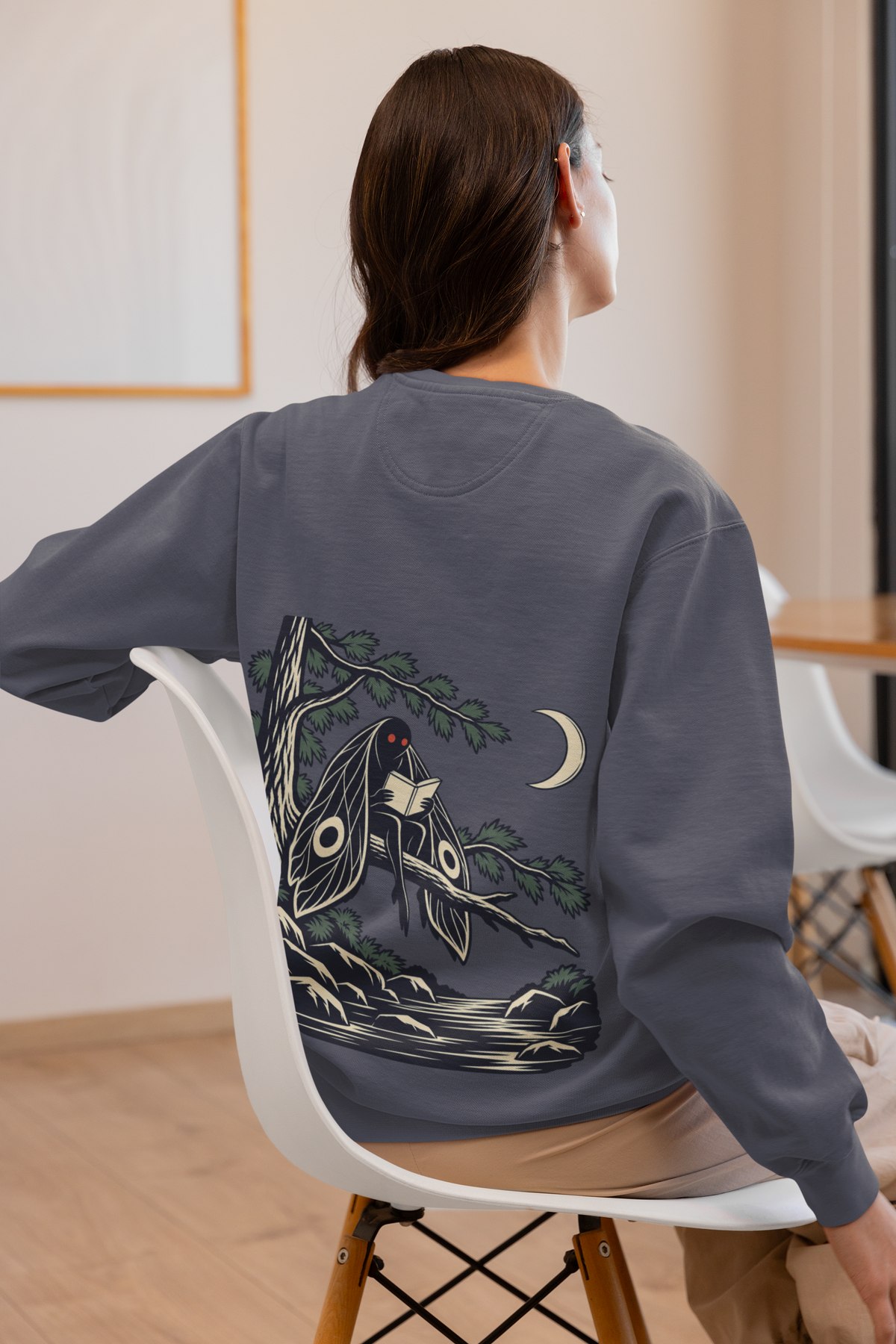 Night Library Garment-Dyed Crewneck in Denim, worn by a woman seen from behind at home, showing the large print on the back. Digital product mockup.