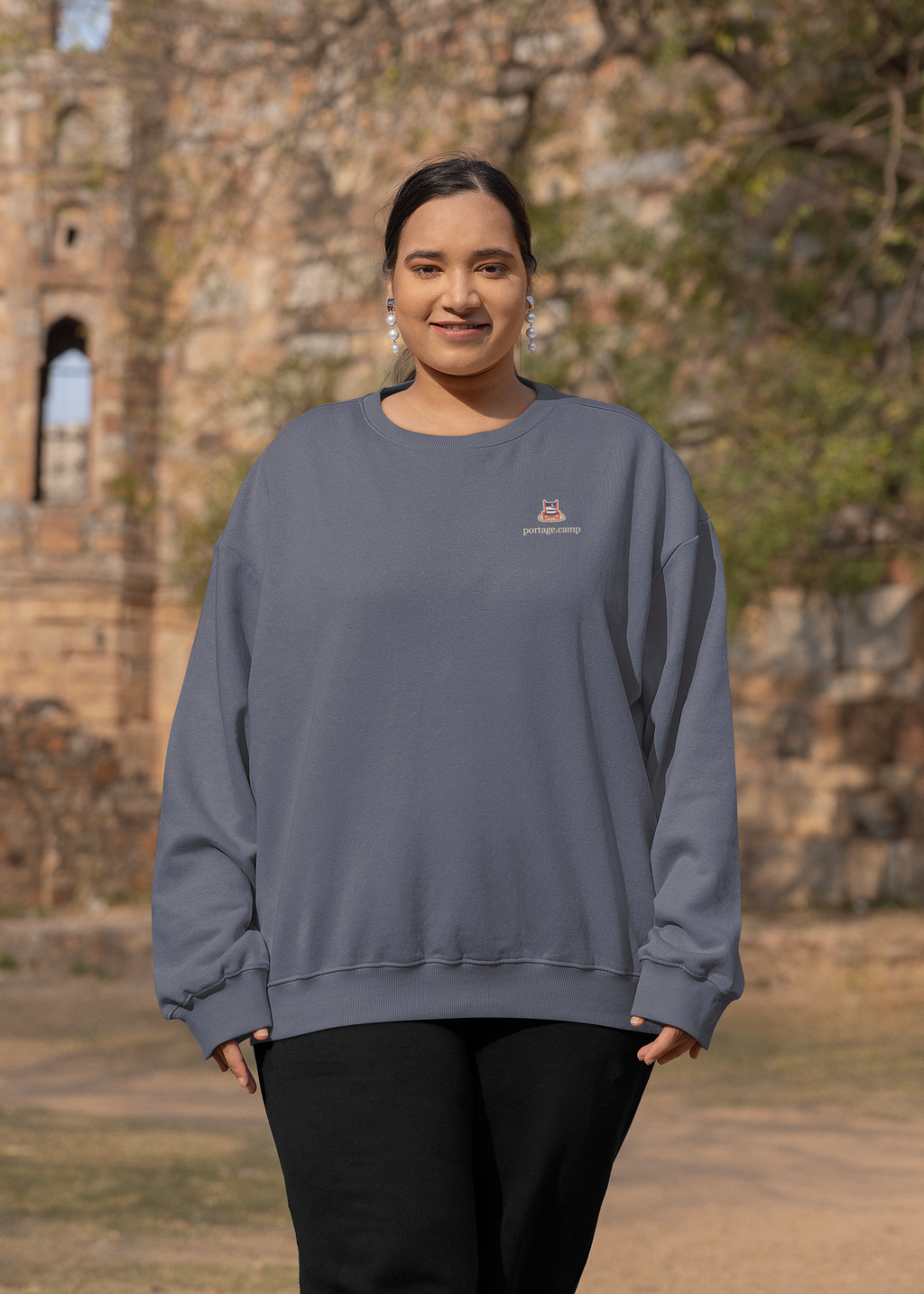 Edge of Evening Garment-Dyed Crewneck in Denim, worn by a woman by an old stone building, with the Edge of Evening logo and portage.camp on the chest. Digital product mockup.