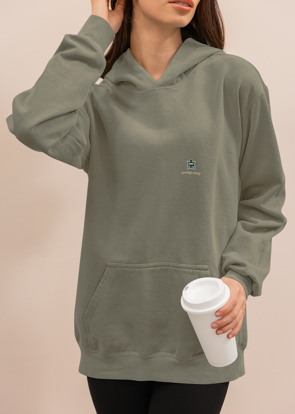 Night Library Heavyweight Hoodie in Olive, worn by a woman at home with a coffee cup, with the Night Library logo and portage.camp on the left chest. Digital product mockup.