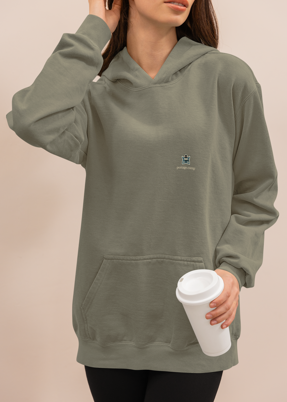 Night Library Heavyweight Hoodie in Olive, worn by a woman at home with a coffee cup, with the Night Library logo and portage.camp on the left chest. Digital product mockup.