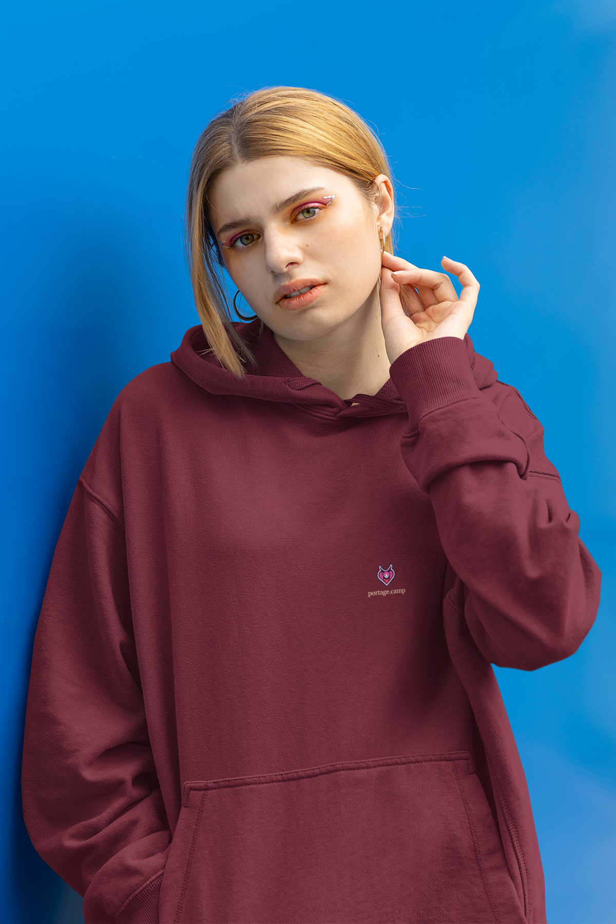 Heart Thief Midweight Hoodie in Maroon, worn by a woman against a bright blue wall, with the Heart Thief logo and portage.camp on the chest. Digital product mockup.