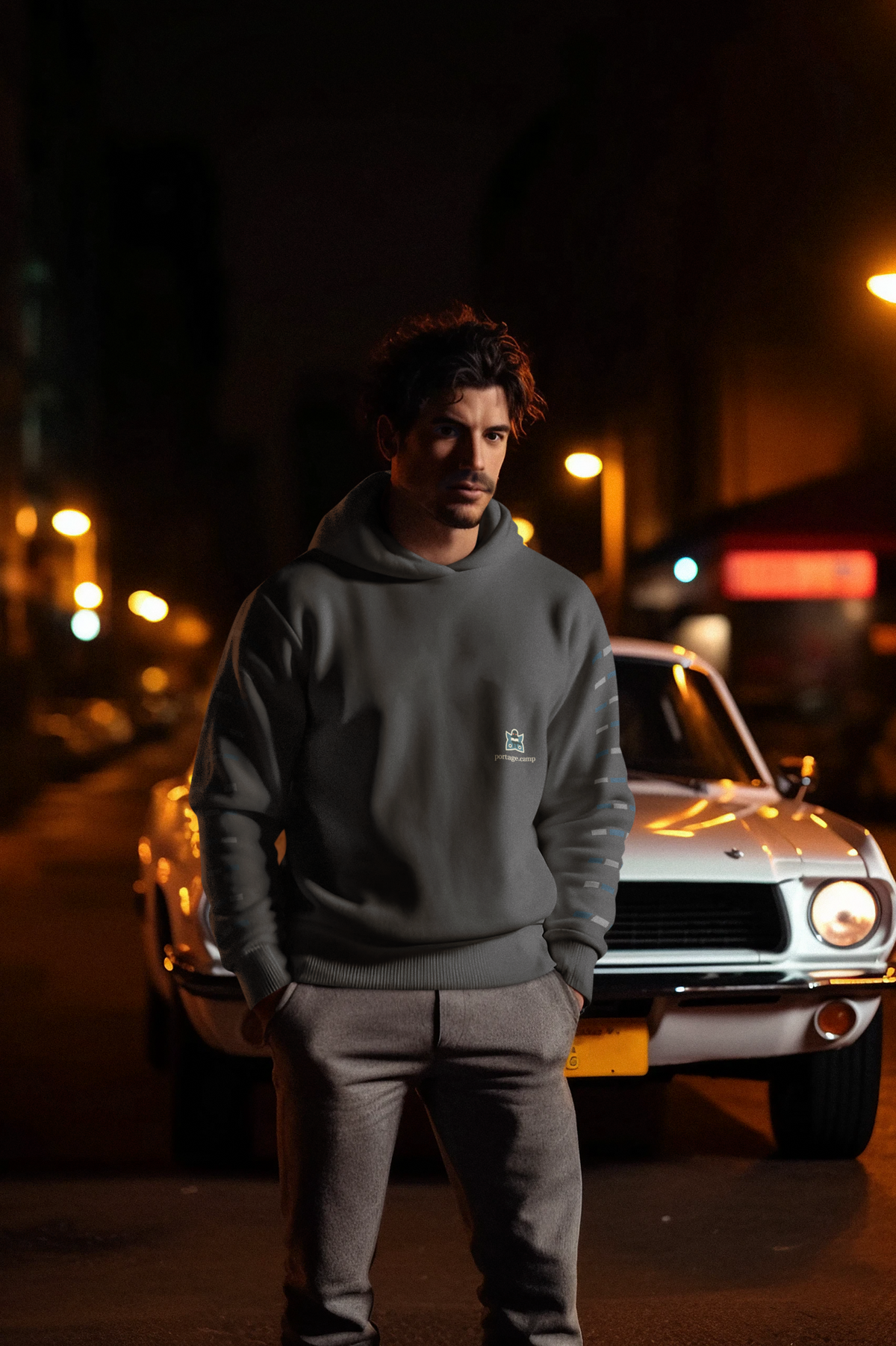 Night Library Heavyweight Hoodie in Black, worn by a man standing by a classic car at night, with the Night Library logo and portage.camp on the left chest. Digital product mockup.