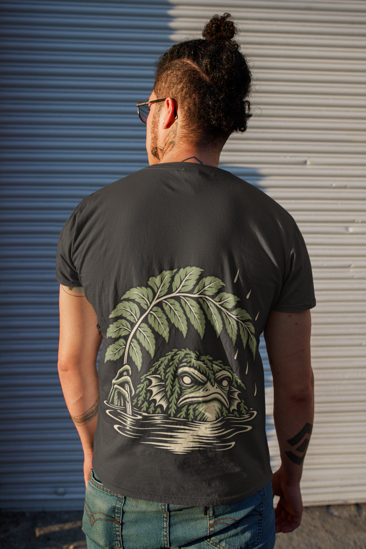 Rain Check Garment-Dyed Tee in Black, worn by a man seen from behind on a street beside a metal shutter, showing the large print on the back. Digital product mockup.