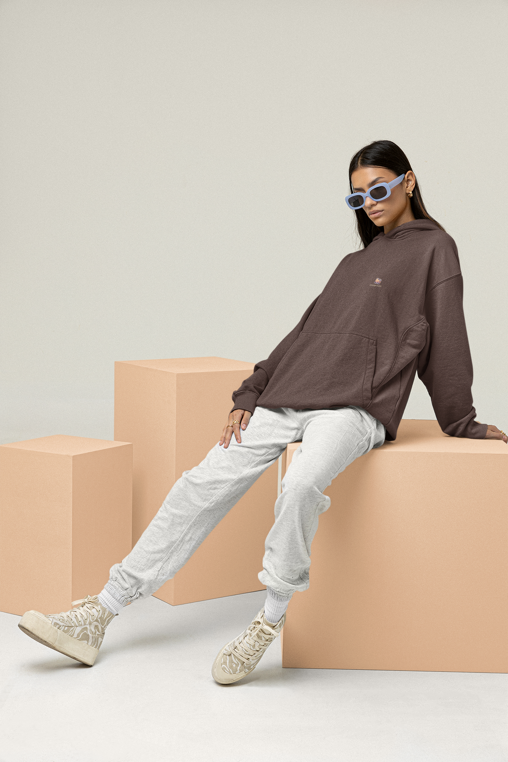 Stay for the Sky Heavyweight Hoodie in Brown, worn by a woman leaning back on studio blocks, with the Stay for the Sky logo and portage.camp on the left chest. Digital product mockup.