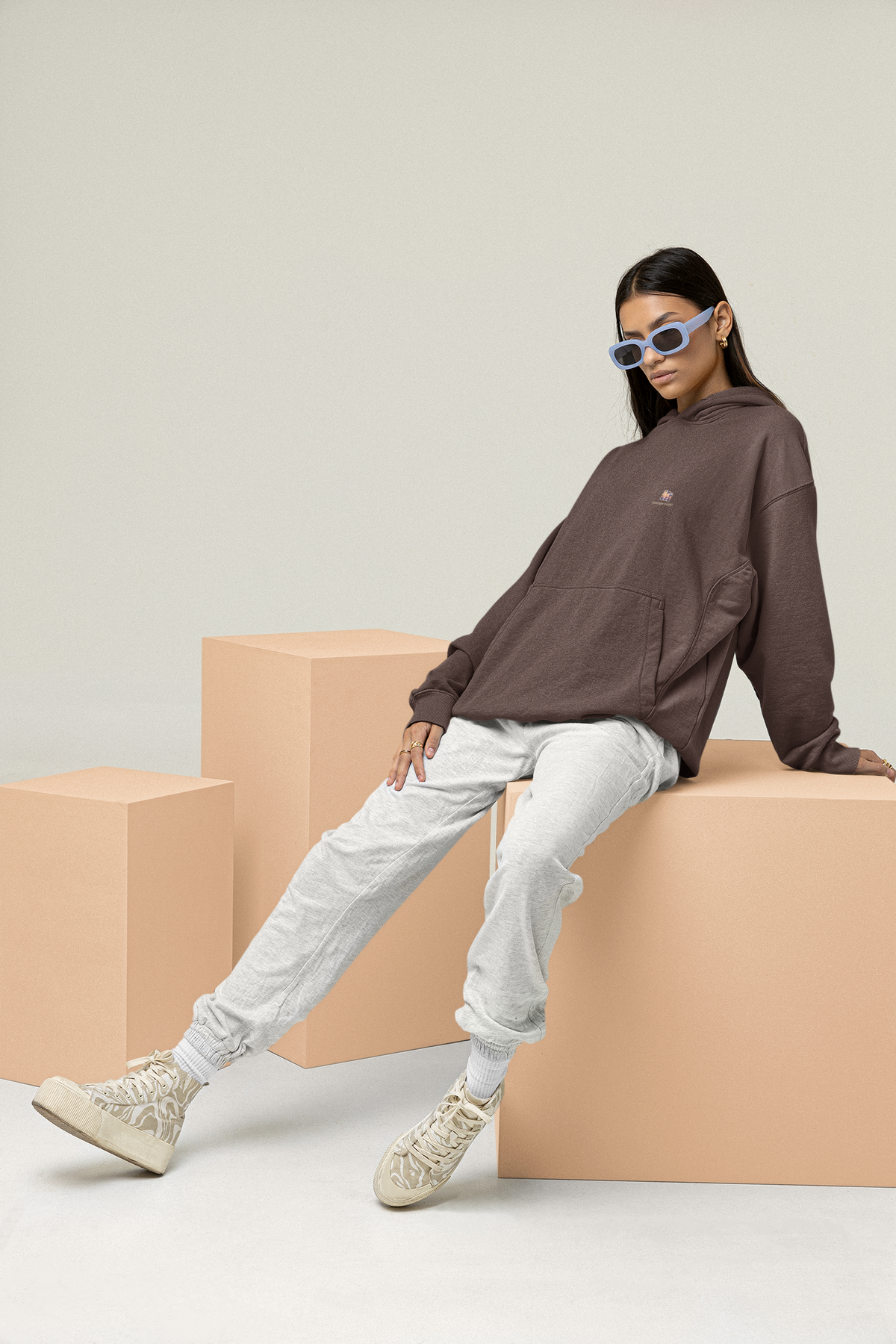 Stay for the Sky Heavyweight Hoodie in Brown, worn by a woman leaning back on studio blocks, with the Stay for the Sky logo and portage.camp on the left chest. Digital product mockup.