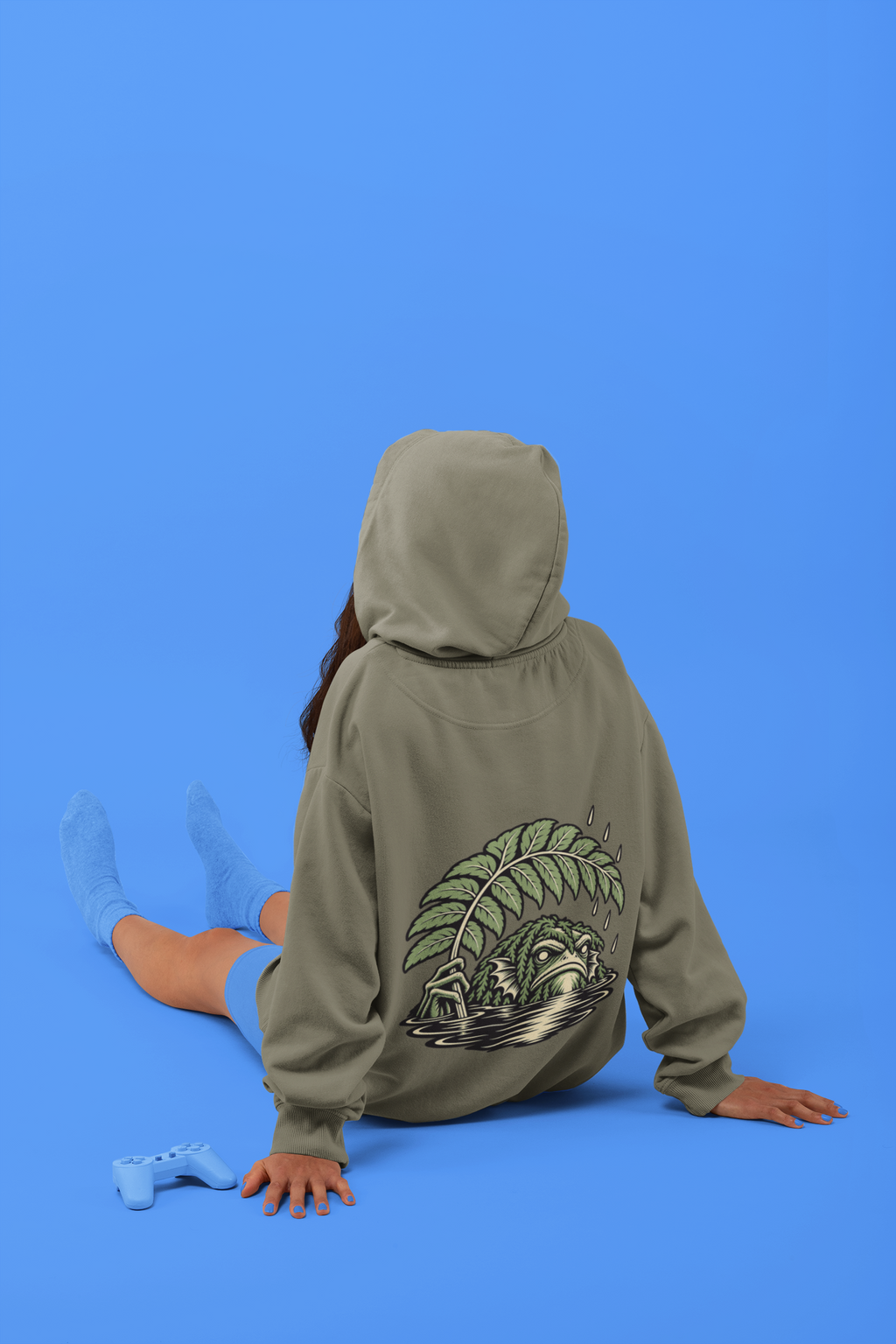 Rain Check Heavyweight Hoodie in Olive, worn by a woman seen from behind seated on a studio floor, showing the large print on the back. Digital product mockup.