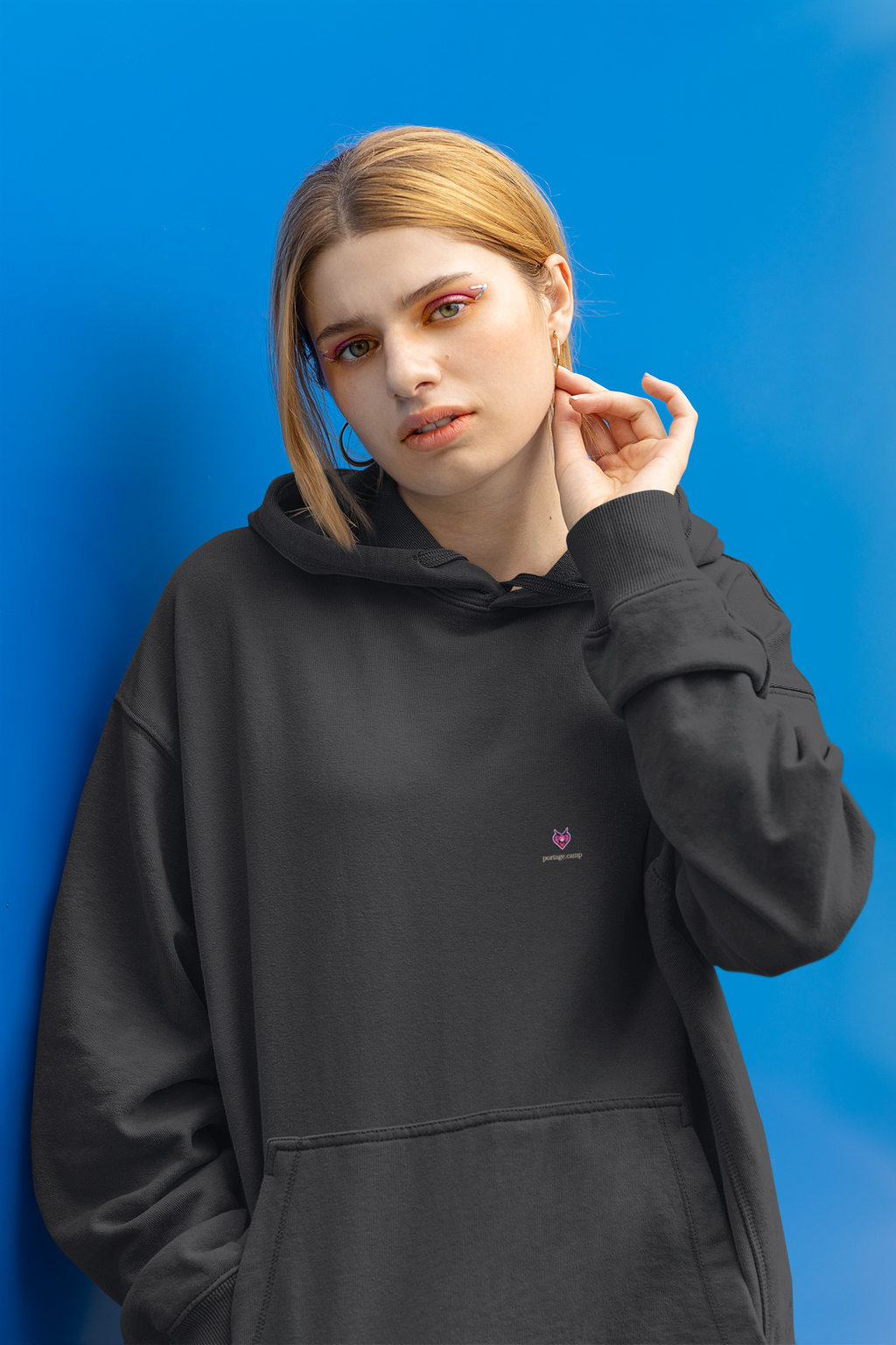 Heart Thief Heavyweight Hoodie in Black, worn by a woman against a bright blue wall, with the Heart Thief logo and portage.camp on the left chest. Digital product mockup.
