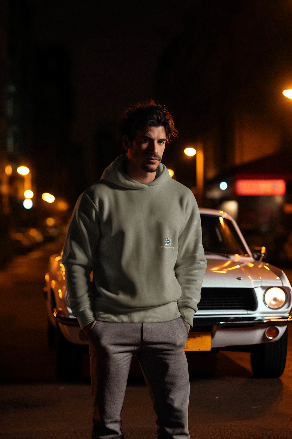 Night Library Heavyweight Hoodie in Olive, worn by a man standing by a classic car at night, with the Night Library logo and portage.camp on the left chest. Digital product mockup.