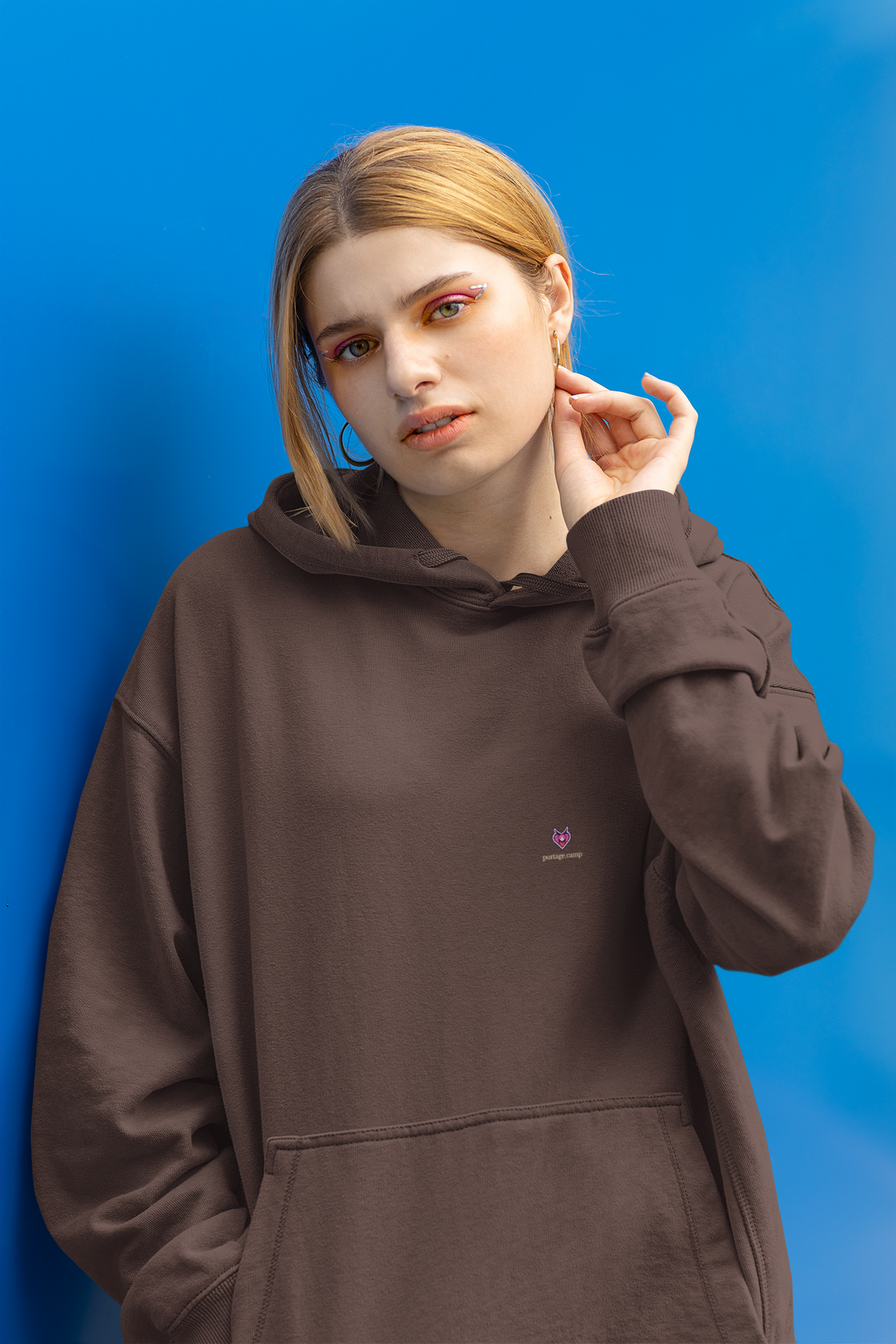 Heart Thief Heavyweight Hoodie in Brown, worn by a woman against a bright blue wall, with the Heart Thief logo and portage.camp on the left chest. Digital product mockup.