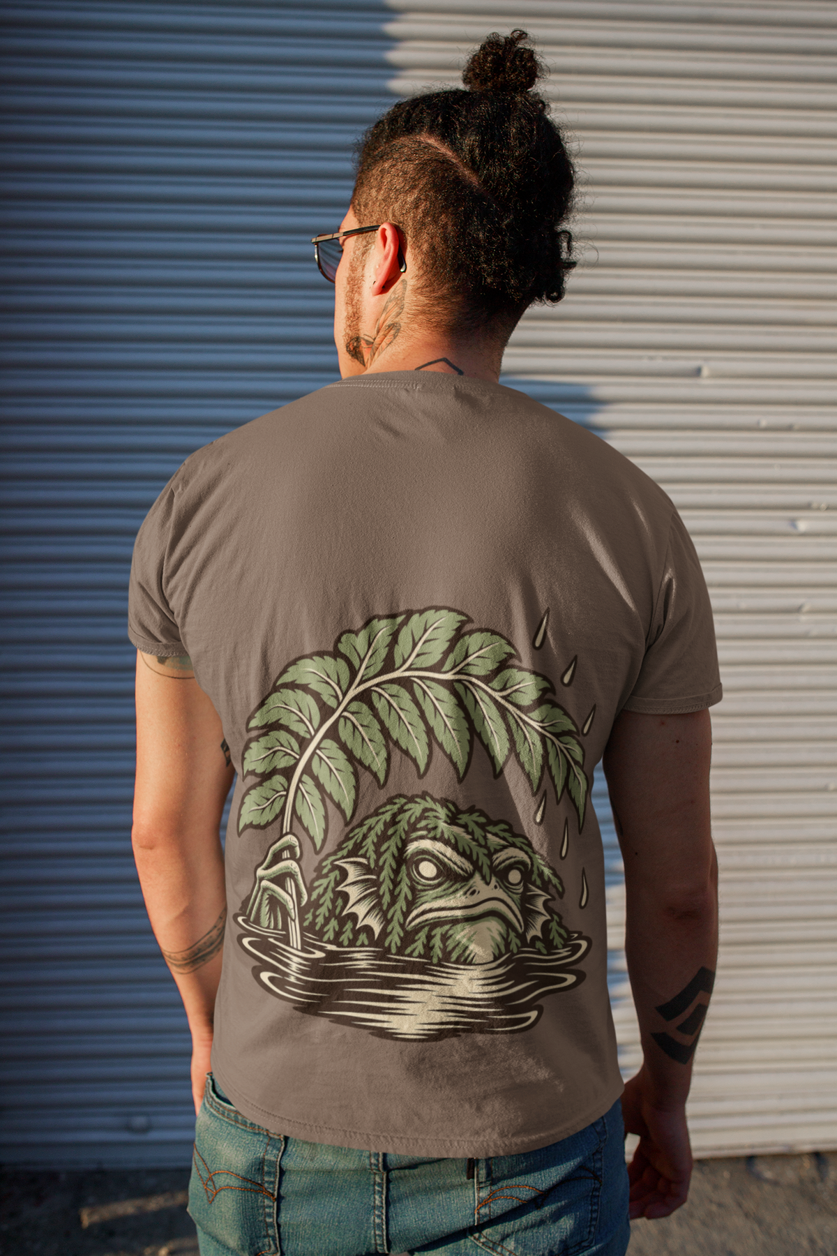Rain Check Garment-Dyed Tee in Espresso, worn by a man seen from behind on a street beside a metal shutter, showing the large print on the back. Digital product mockup.