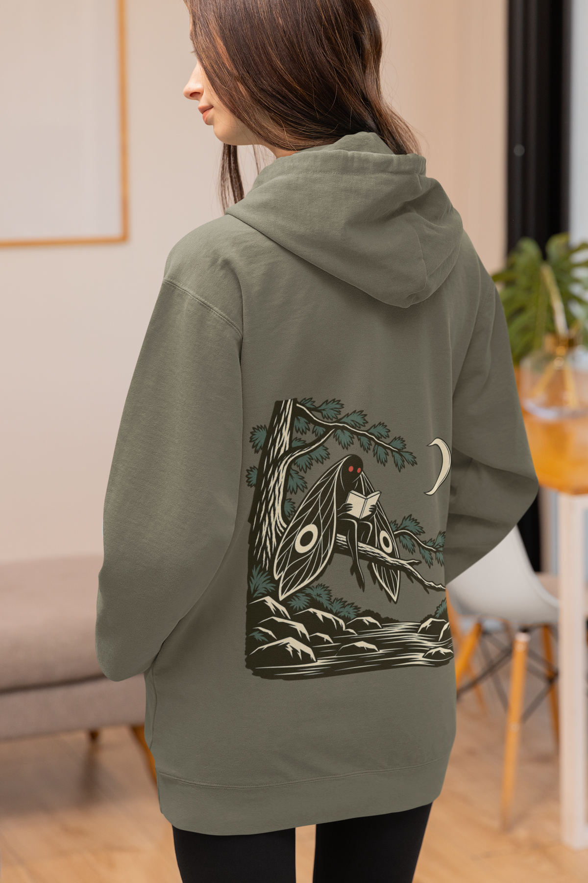 Night Library Heavyweight Hoodie in Olive, worn by a woman seen from behind at home, showing the large print on the back. Digital product mockup.