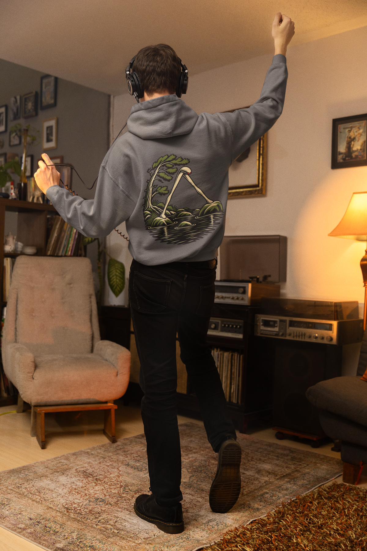 Small Hours Midweight Hoodie in Charcoal, worn by a man seen from behind in a living room at night, showing the large print on the back. Digital product mockup.