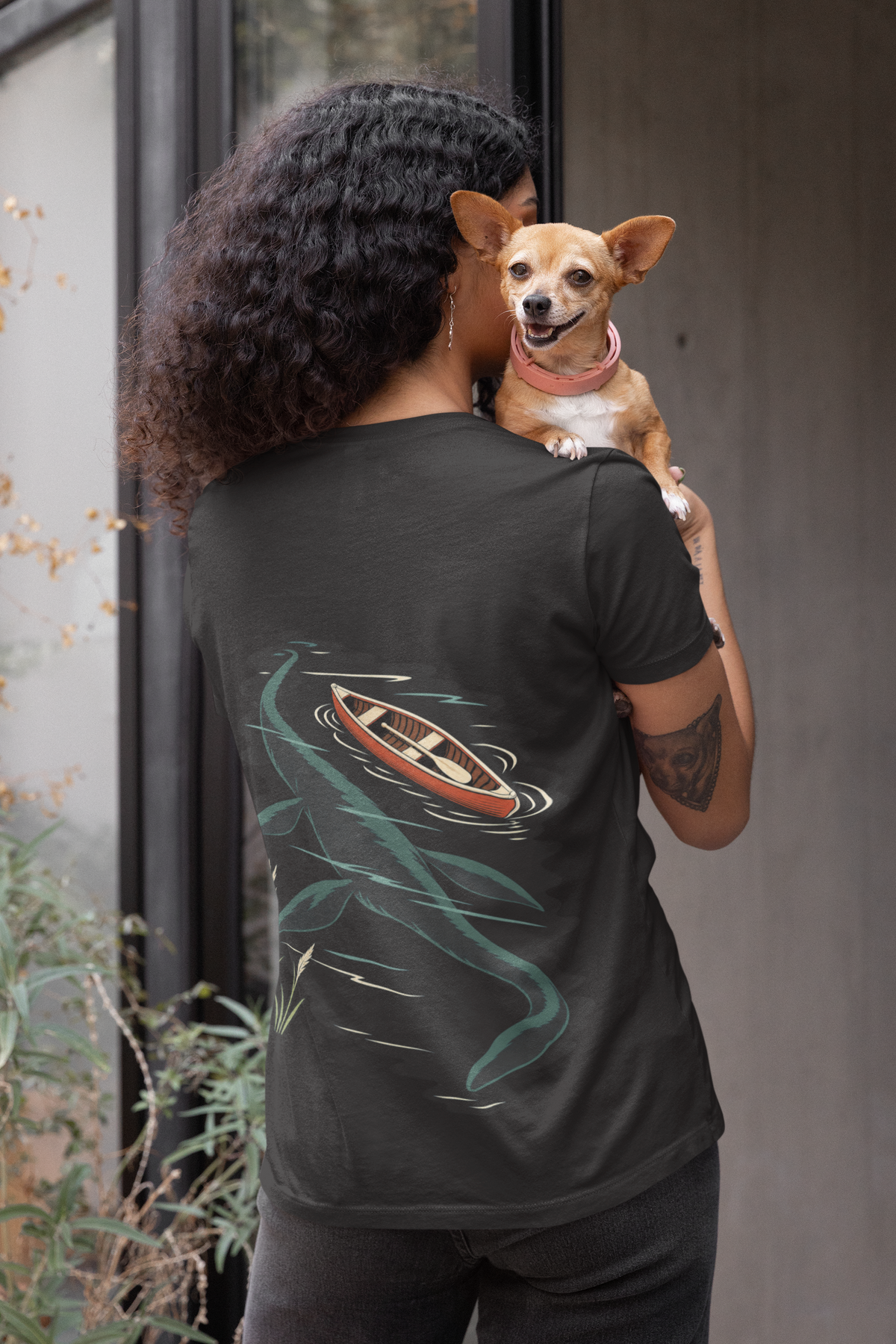 Seen Briefly Garment-Dyed Tee in Black, worn by a woman seen from behind holding a small dog, showing the large print on the back. Digital product mockup.