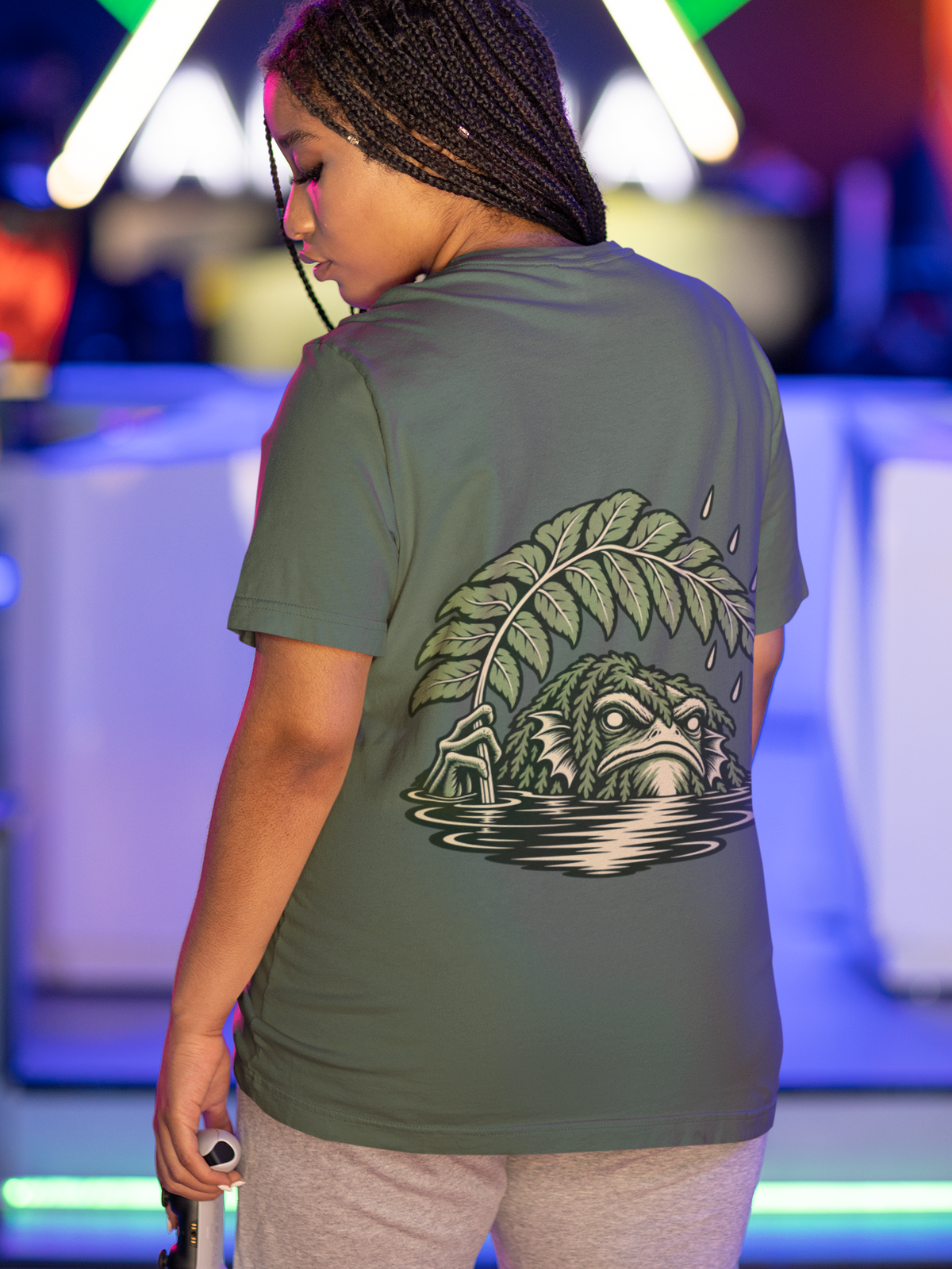 Rain Check Garment-Dyed Tee in Blue Spruce, worn by a woman seen from behind under neon lights in a gaming arcade, showing the large print on the back. Digital product mockup.
