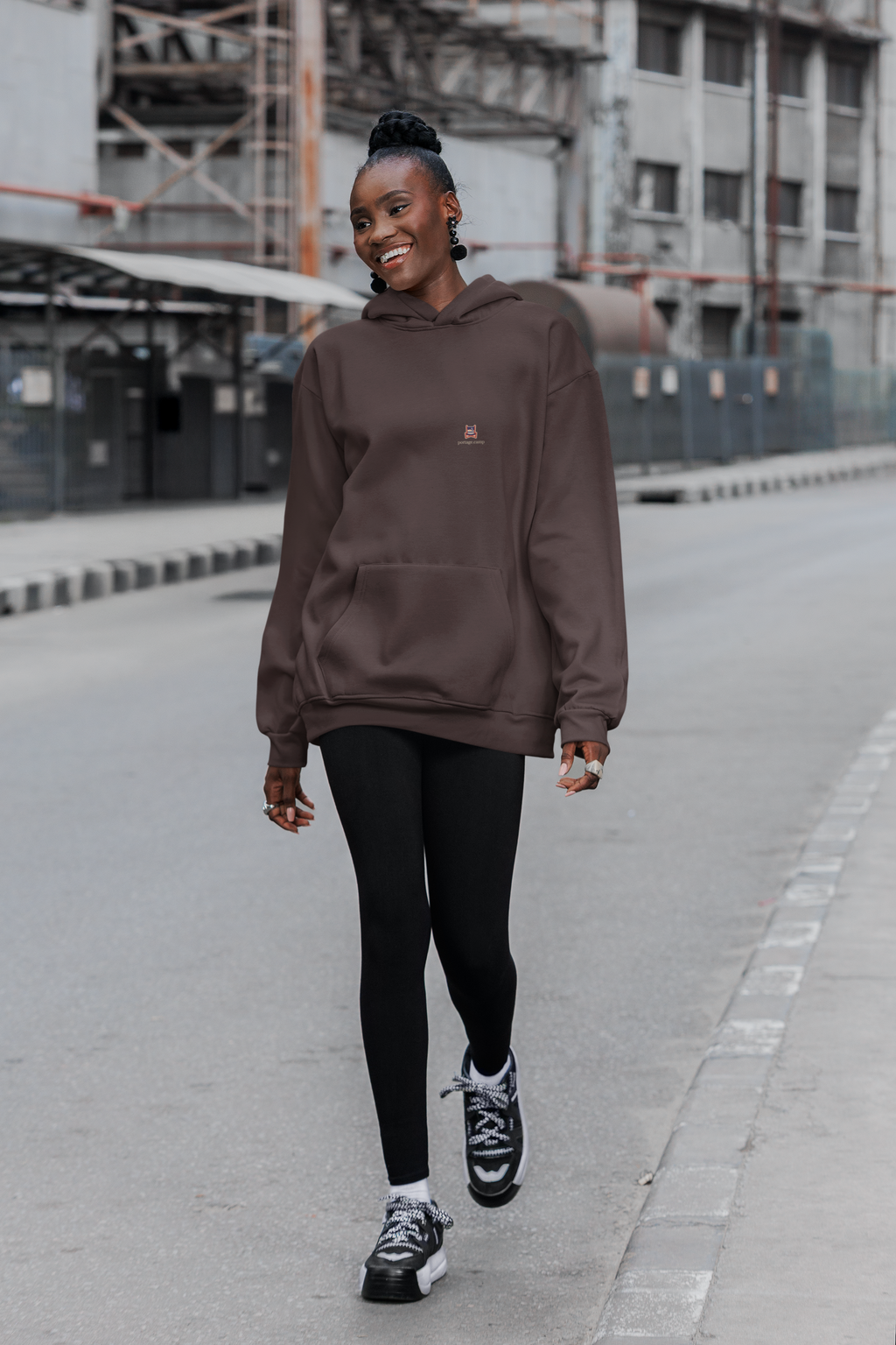 Edge of Evening Heavyweight Hoodie in Brown, worn by a woman walking down a city street, with the Edge of Evening logo and portage.camp on the left chest. Digital product mockup.