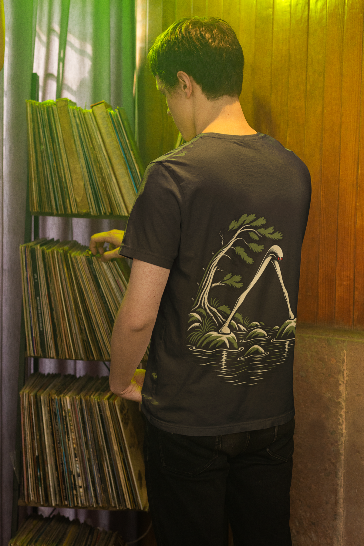 Small Hours Garment-Dyed Tee in Black, worn by a man seen from behind browsing a record shop, showing the large print on the back. Digital product mockup.
