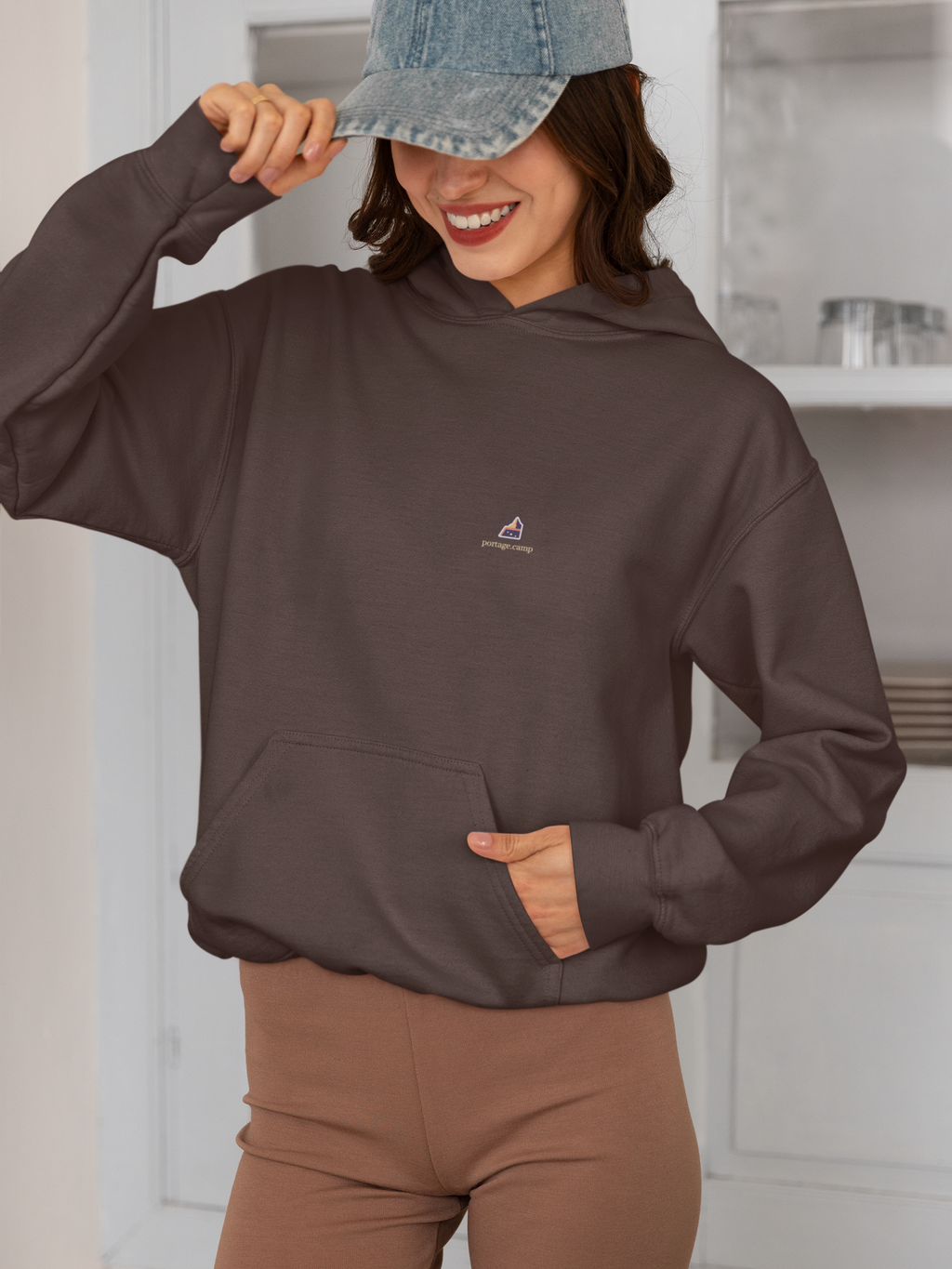 A Different Sky Heavyweight Hoodie in Brown, worn by a woman smiling in a kitchen, with the A Different Sky logo and portage.camp on the left chest. Digital product mockup.
