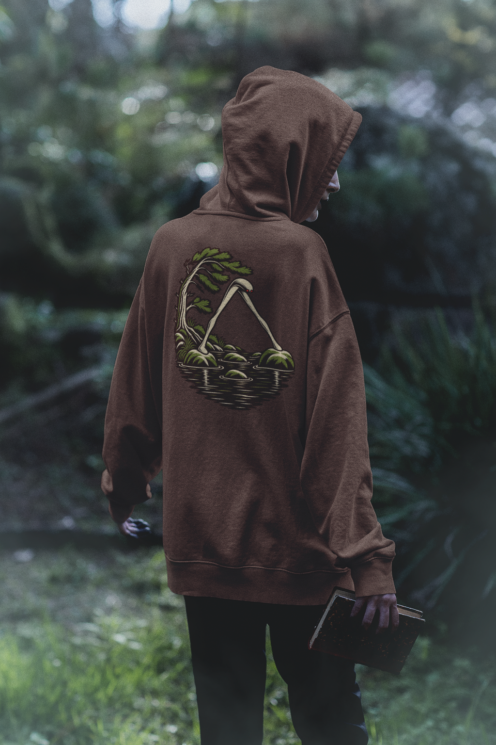 Small Hours Heavyweight Hoodie in Brown, worn by a woman seen from behind in a dark forest, showing the large print on the back. Digital product mockup.