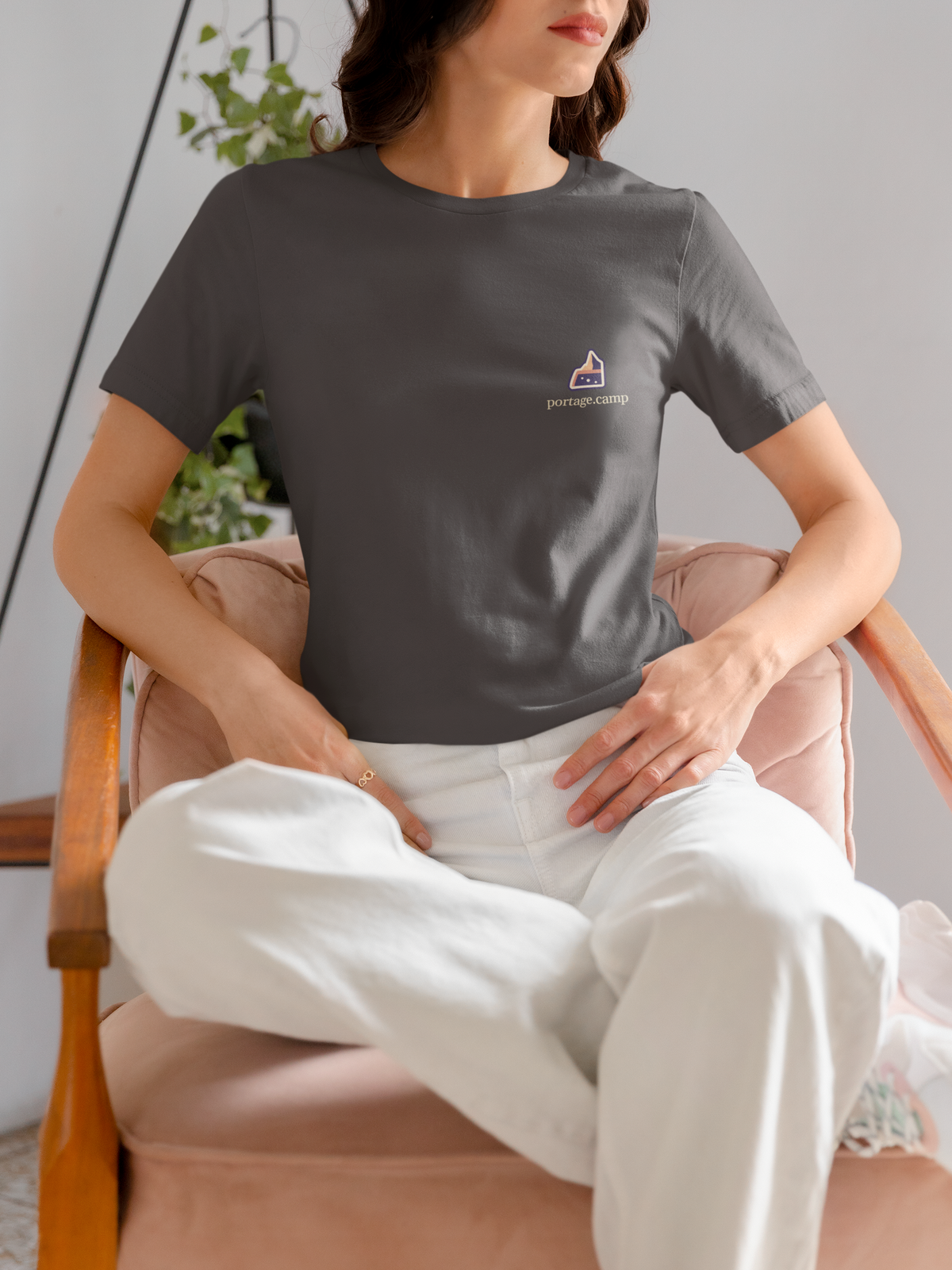 A Different Sky Garment-Dyed Tee in Graphite, worn by a woman seated on a chair at home, with the A Different Sky logo and portage.camp on the chest. Digital product mockup.