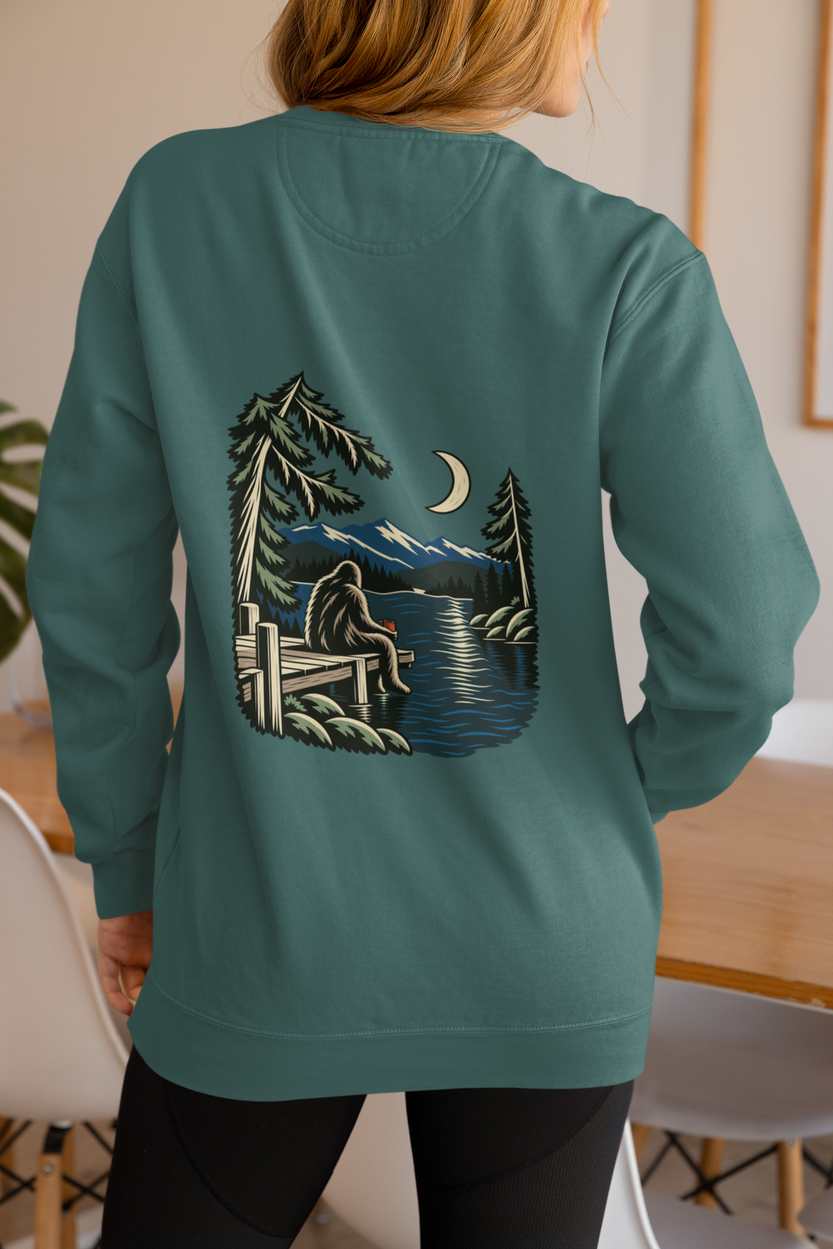 Quiet Hours Garment-Dyed Crewneck in Blue Spruce, worn by a woman seen from behind at home, showing the large print on the back. Digital product mockup.