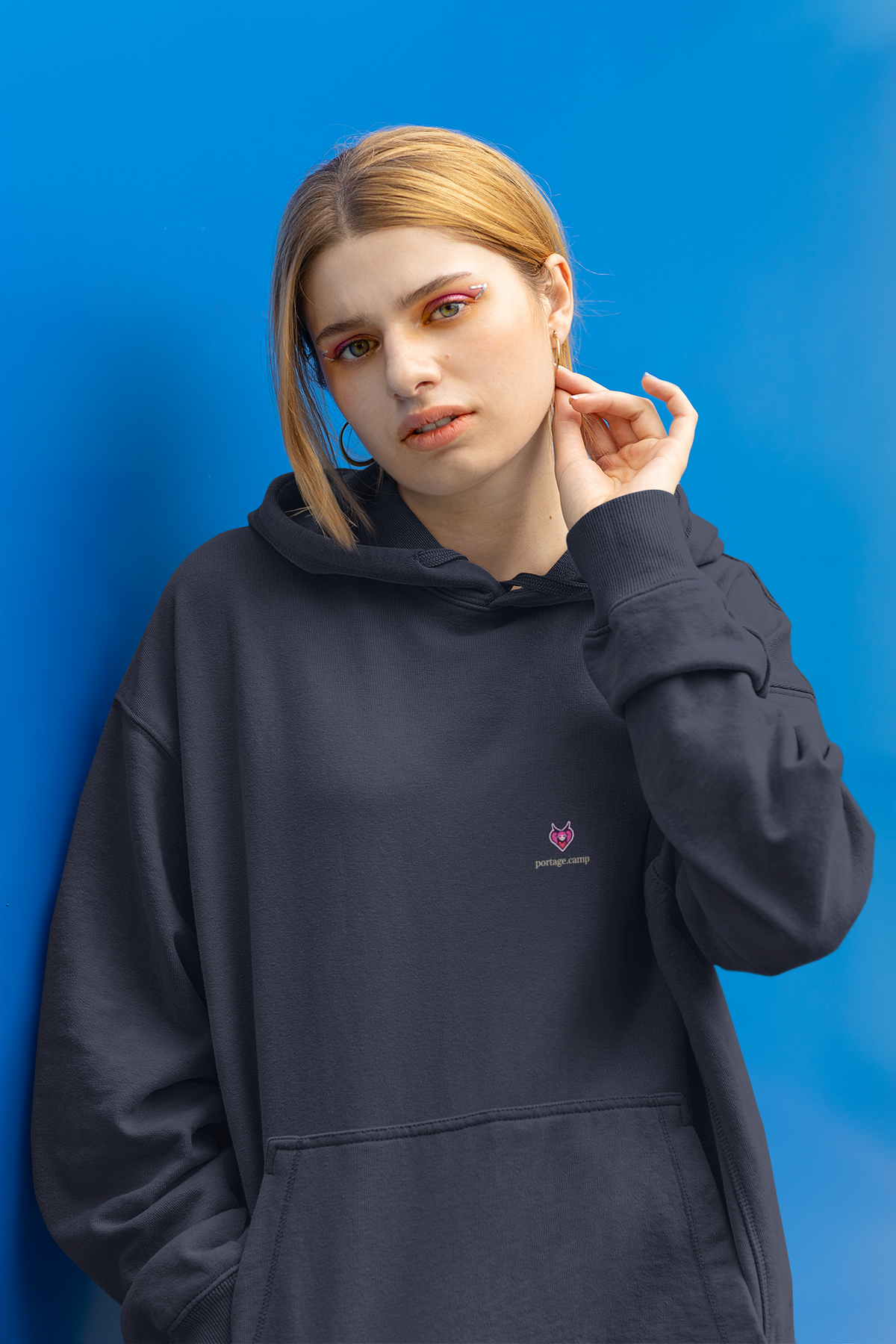 Heart Thief Midweight Hoodie in Classic Navy, worn by a woman against a bright blue wall, with the Heart Thief logo and portage.camp on the chest. Digital product mockup.